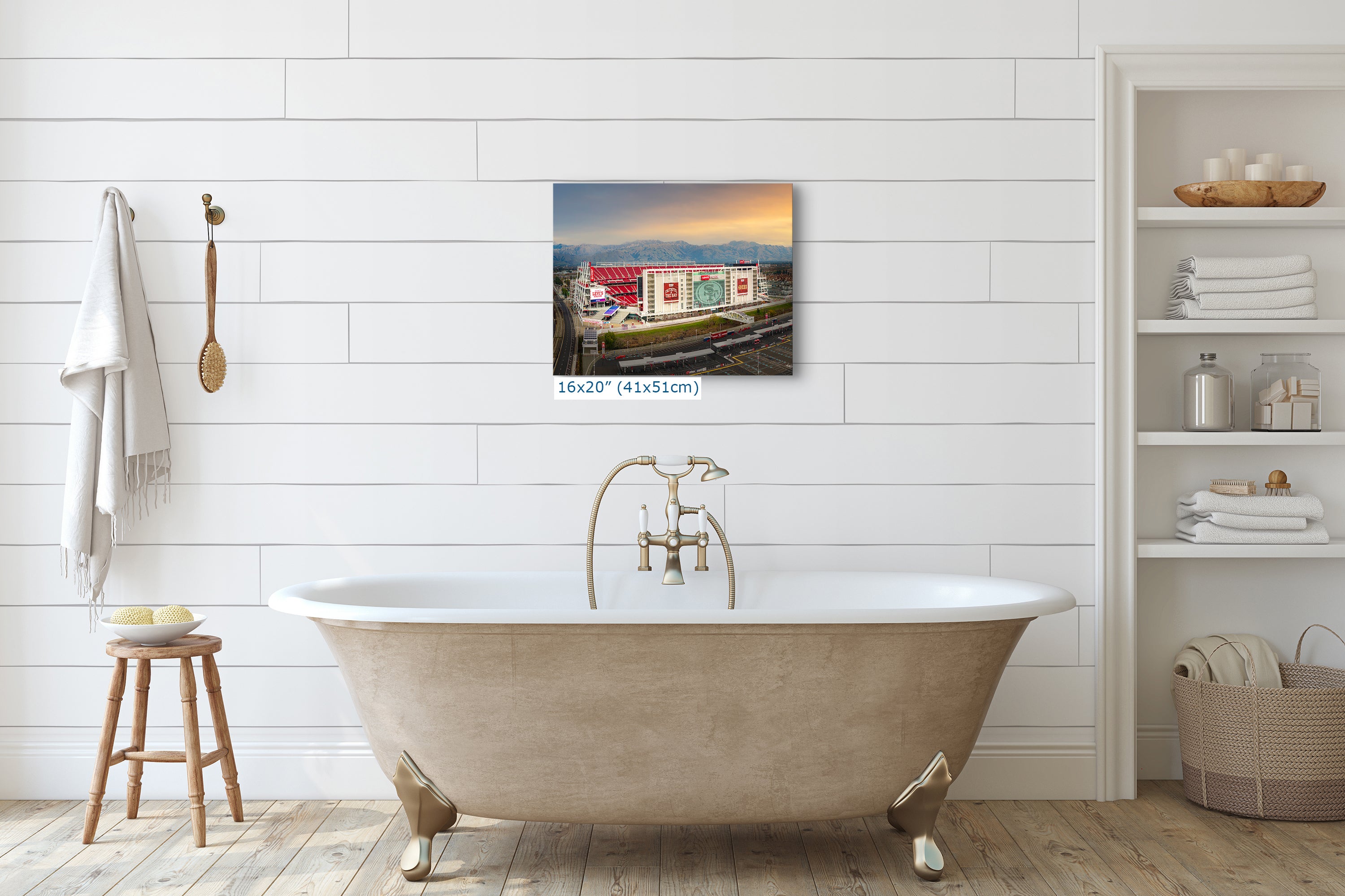 a bathroom with a clawfoot bathtub, a wooden stool, and a shelf with various toiletries. Above the bathtub, there is a framed photograph of a building with a red roof and a white building with a blue roof.