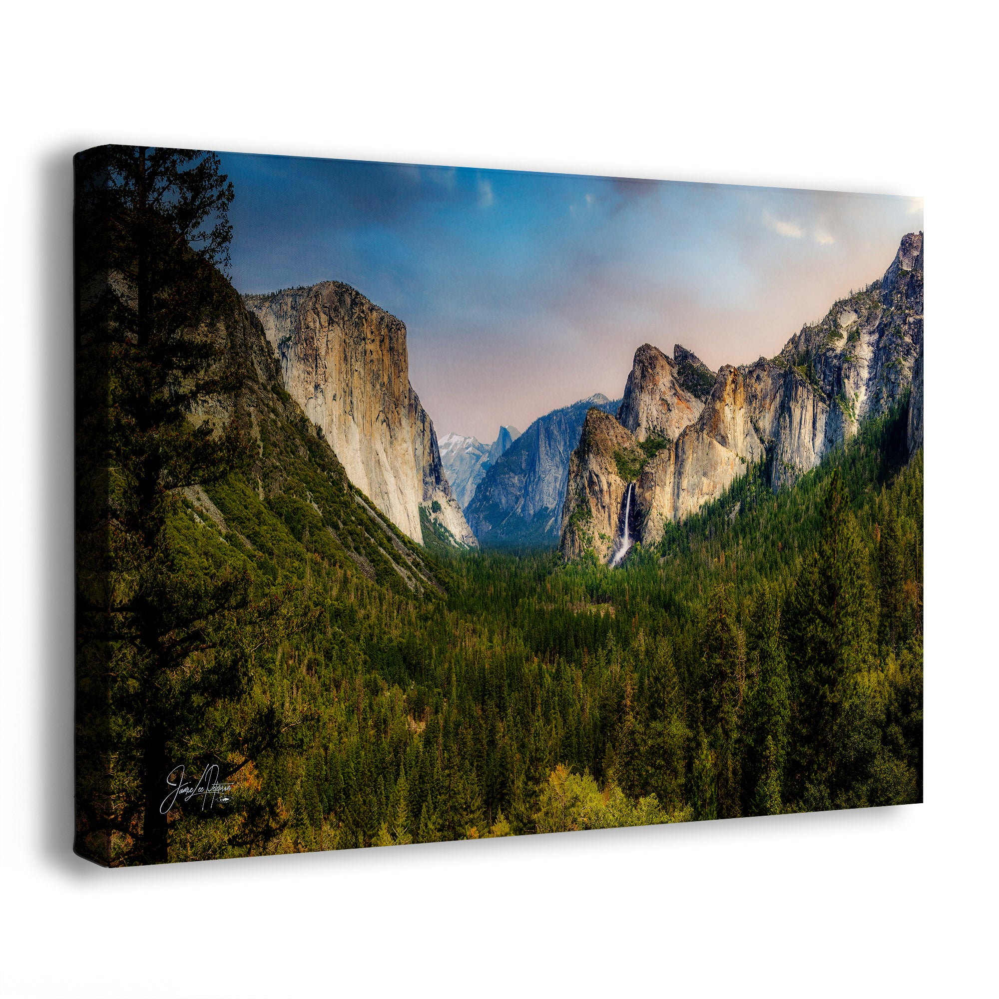 A canvas wall art print showing a dramatic mountain valley landscape with tall rock cliffs, dense evergreen forest, and a blue sky with soft clouds. The canvas is photographed at a slight angle against a clean white background.