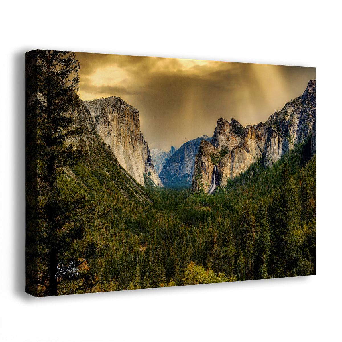 A rectangular canvas wall art print showing a dramatic mountain valley landscape with tall rock cliffs, dense evergreen forest, and golden-brown clouds with sun rays. The canvas is photographed at a slight angle on a clean white background.
