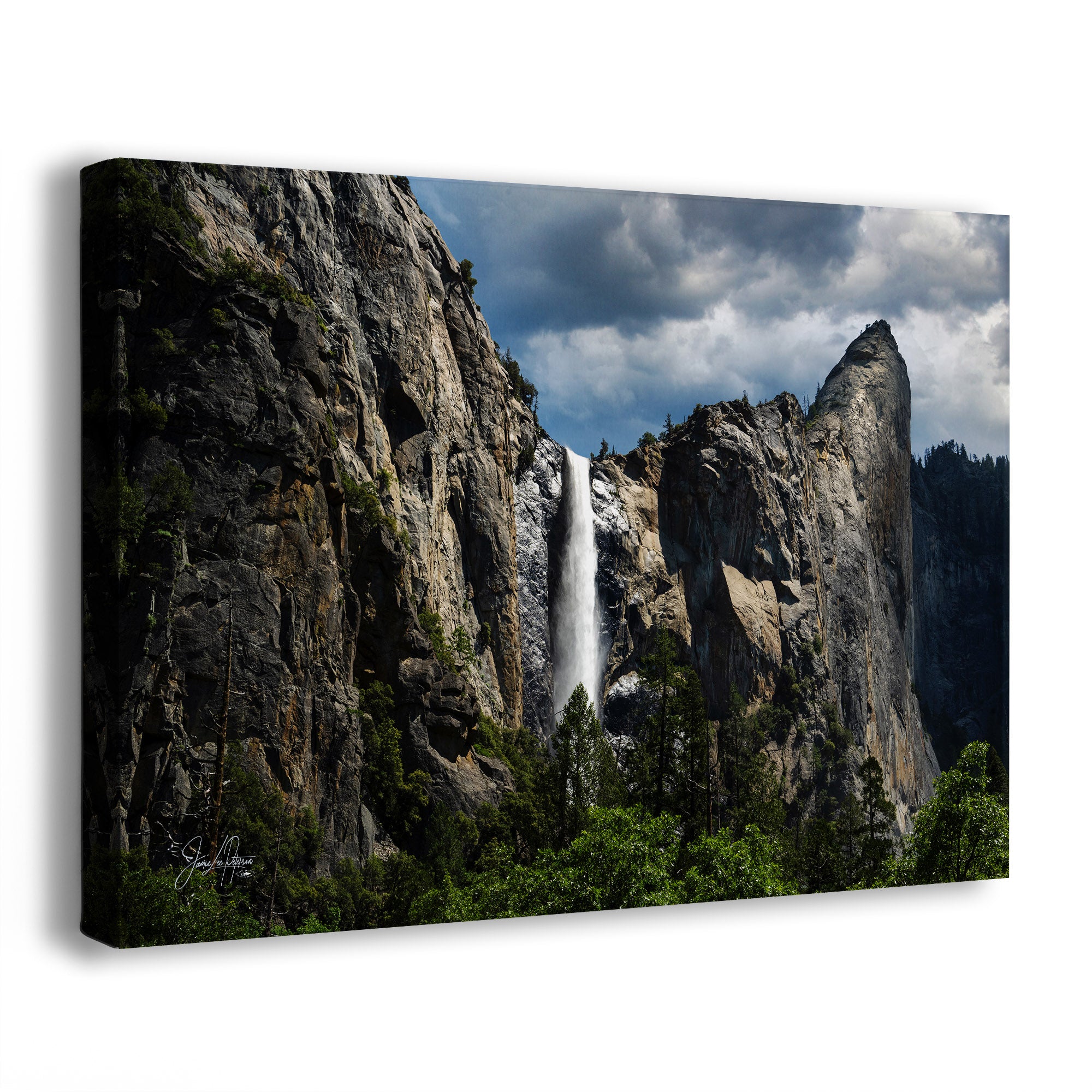 A canvas wall art print showing a dramatic waterfall cascading between tall rocky cliffs under a cloudy blue sky, photographed at an angle against a light gray background.