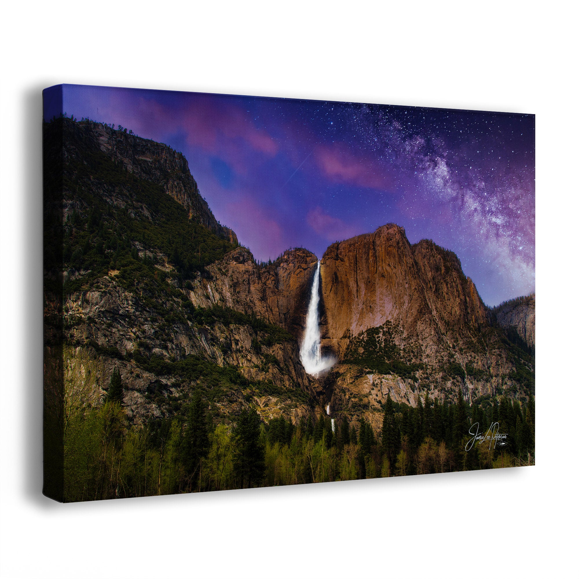 A rectangular canvas wall art print showing a mountain landscape with a tall waterfall, dark evergreen trees, and a dramatic night sky filled with stars and the Milky Way. The canvas is photographed at a slight angle against a clean white background.