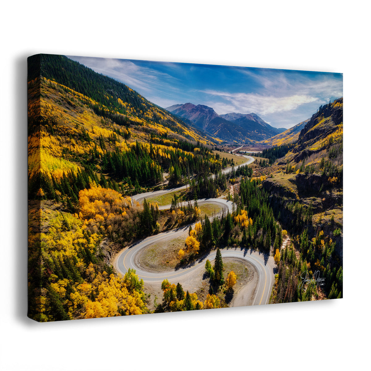 A canvas wall art print showing an aerial Colorado mountain landscape of Million Dollar Highway with a winding road, yellow autumn trees, evergreen forest, and blue sky, photographed against a light gray background.