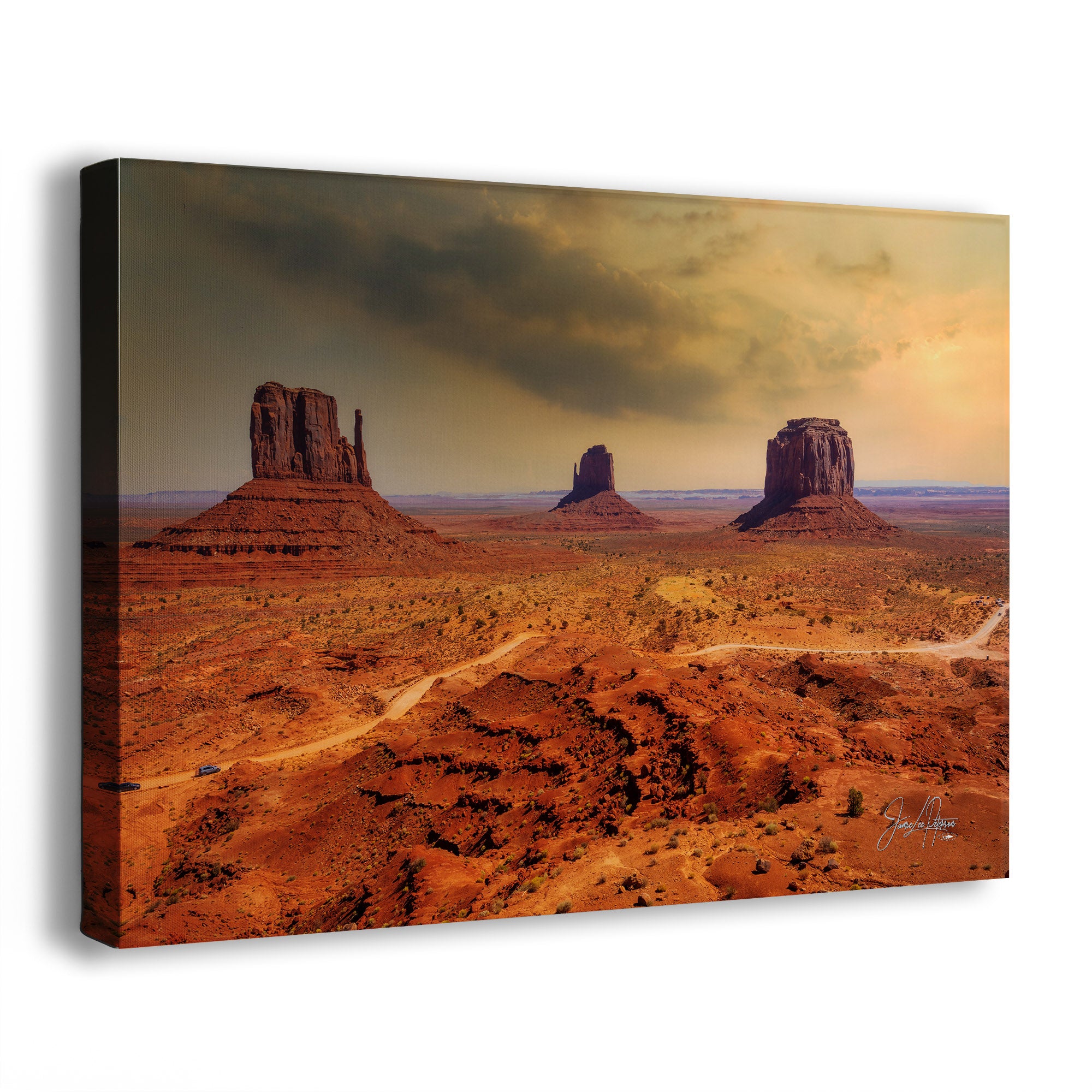 A rectangular canvas wall art print showing Monument Valley with red rock buttes under a dramatic cloudy sky, photographed at a slight angle on a white background.