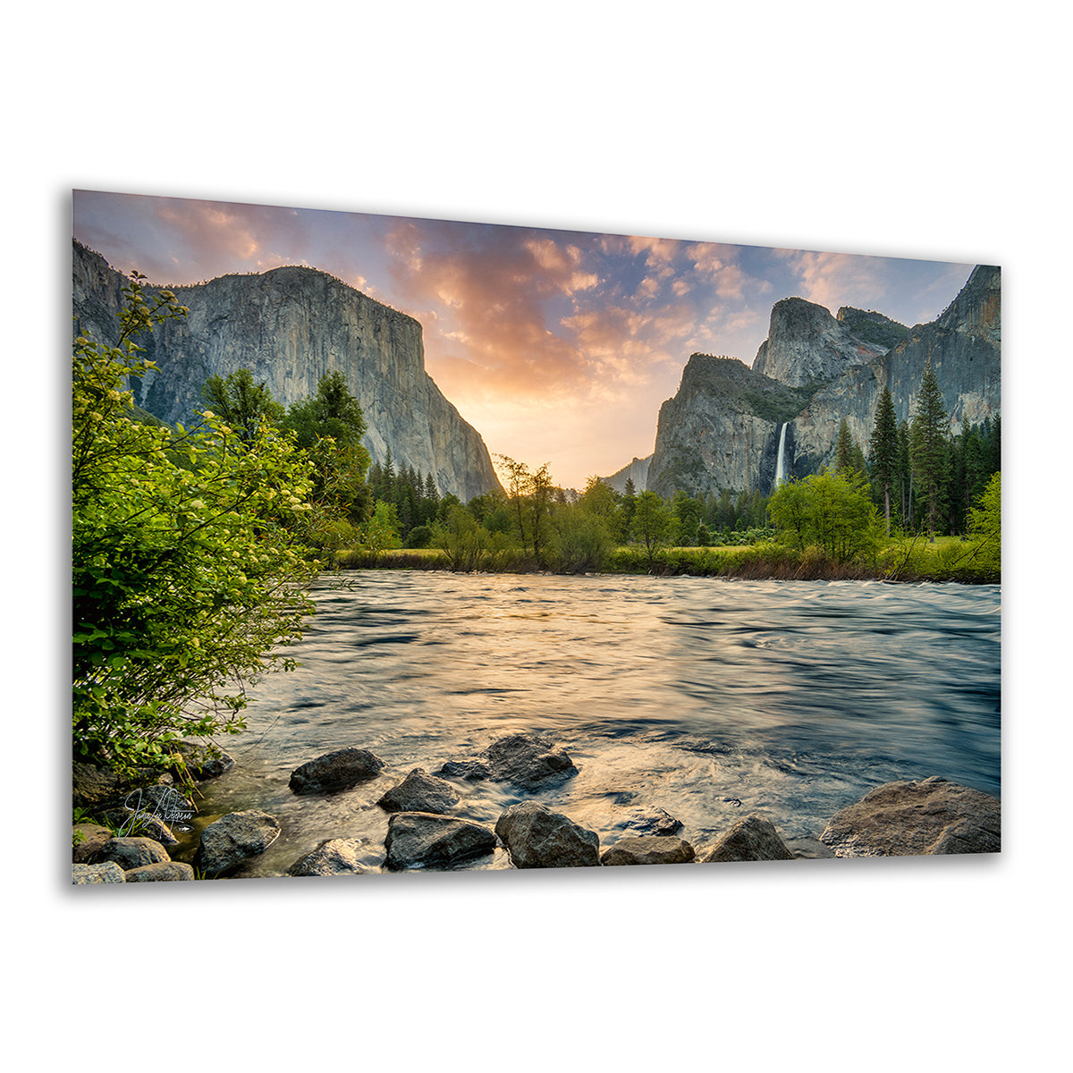 A wall art print or canvas showing a scenic Yosemite mountain landscape with a river, rocks, trees, and a waterfall at sunset, displayed against a plain white background.