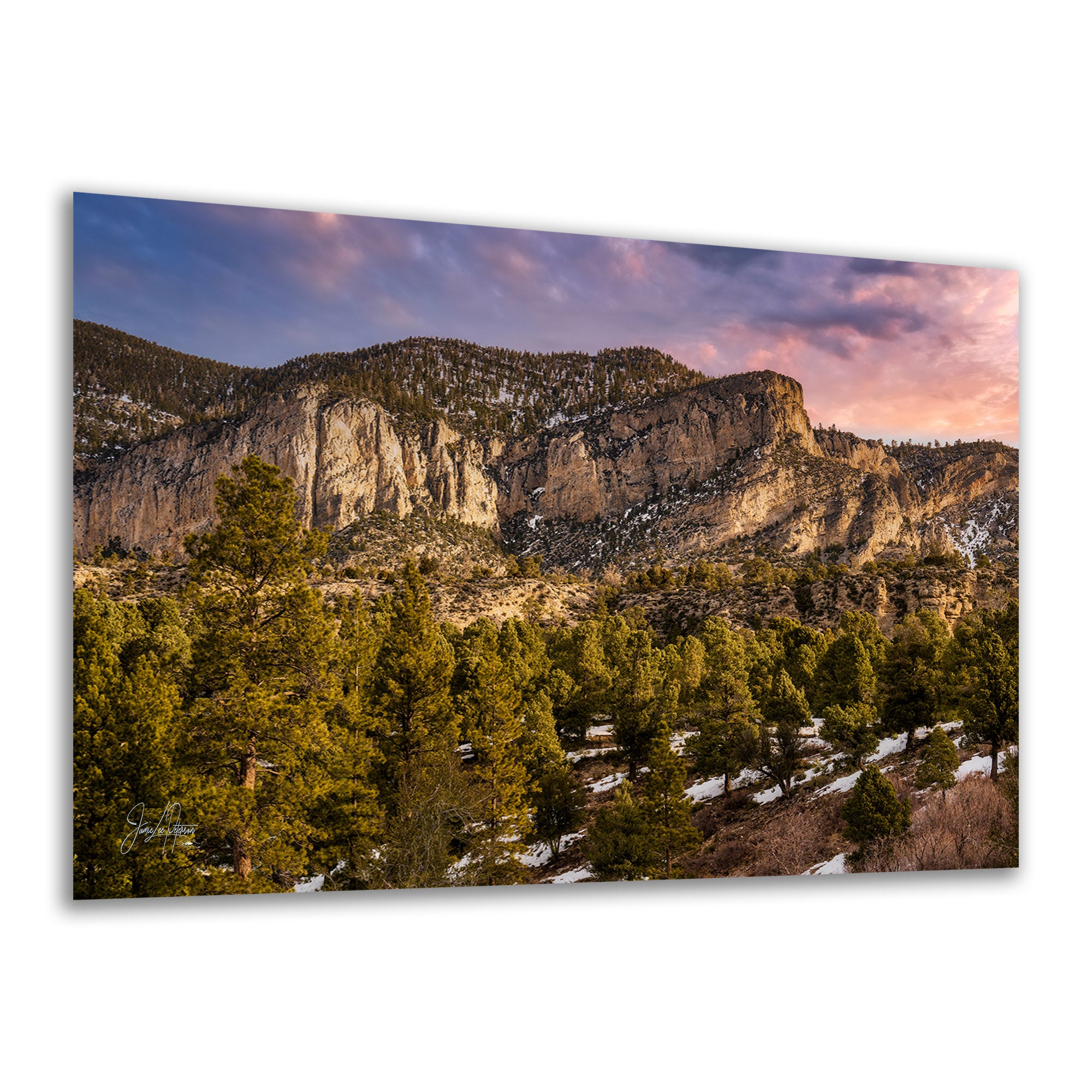 A wall art print showing a Nevada mountain landscape with rocky cliffs, evergreen trees, patches of snow, and a pink-purple sky, photographed against a plain white background.