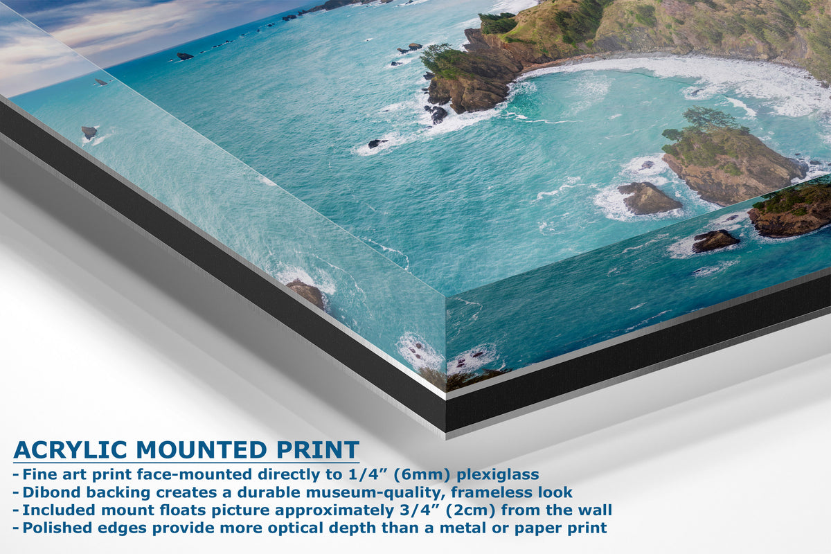 A corner view of an acrylic-mounted art print of a coastal ocean scene with turquoise water and green cliffs, shown at an angle with product text describing frameless mounting and acrylic plexiglass finish.