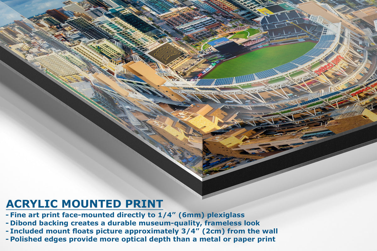 A corner view of an acrylic-mounted art print showing an aerial view of a stadium and surrounding urban area. Blue text at the bottom reads 'ACRYLIC MOUNTED PRINT' with feature bullet points about plexiglass, backing, frameless look, and mount float distance.