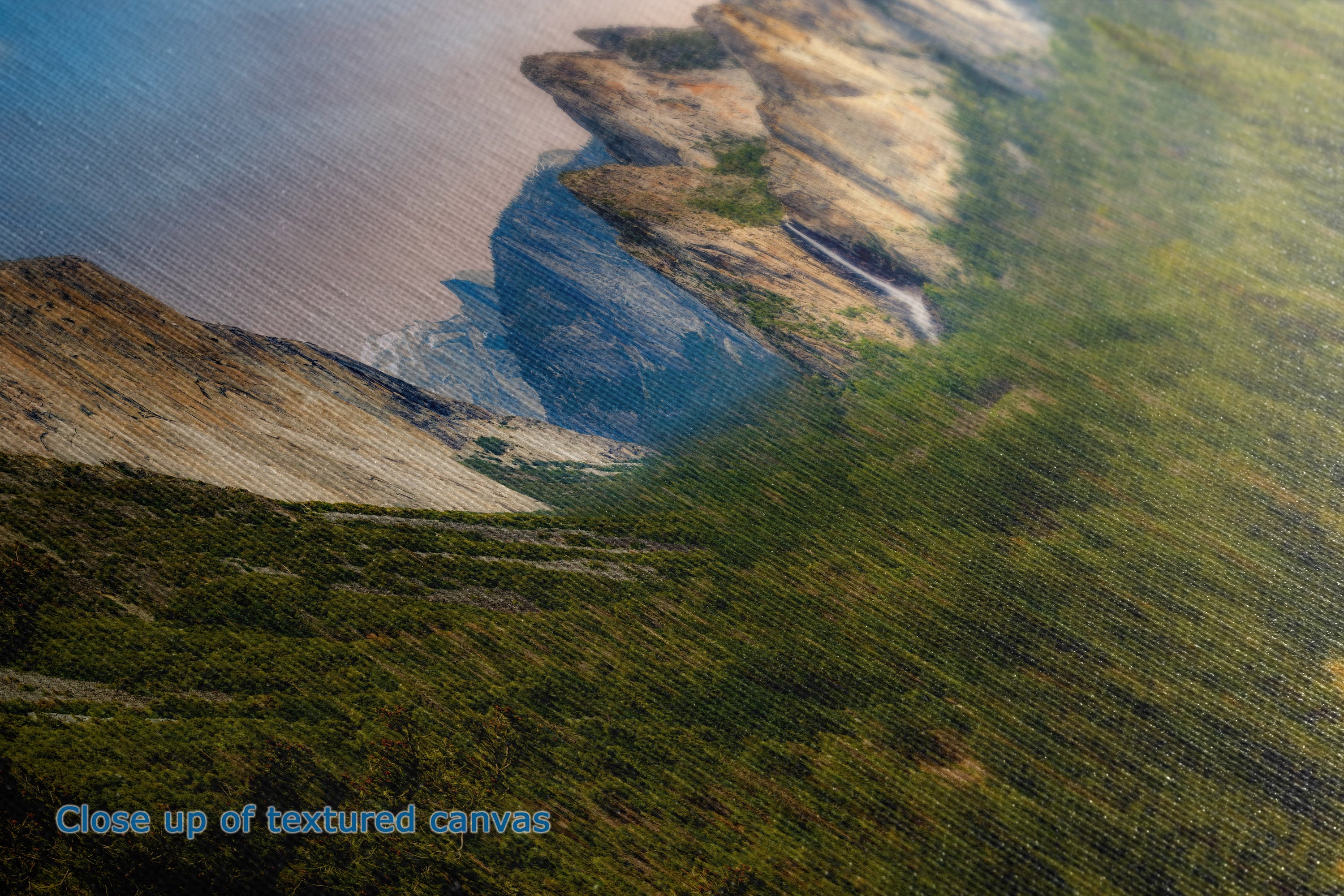 Close-up of a textured canvas print depicting a coastal landscape with green hills, brown cliffs, and blue water. Text in the bottom-left corner reads 'Close up of textured canvases'.