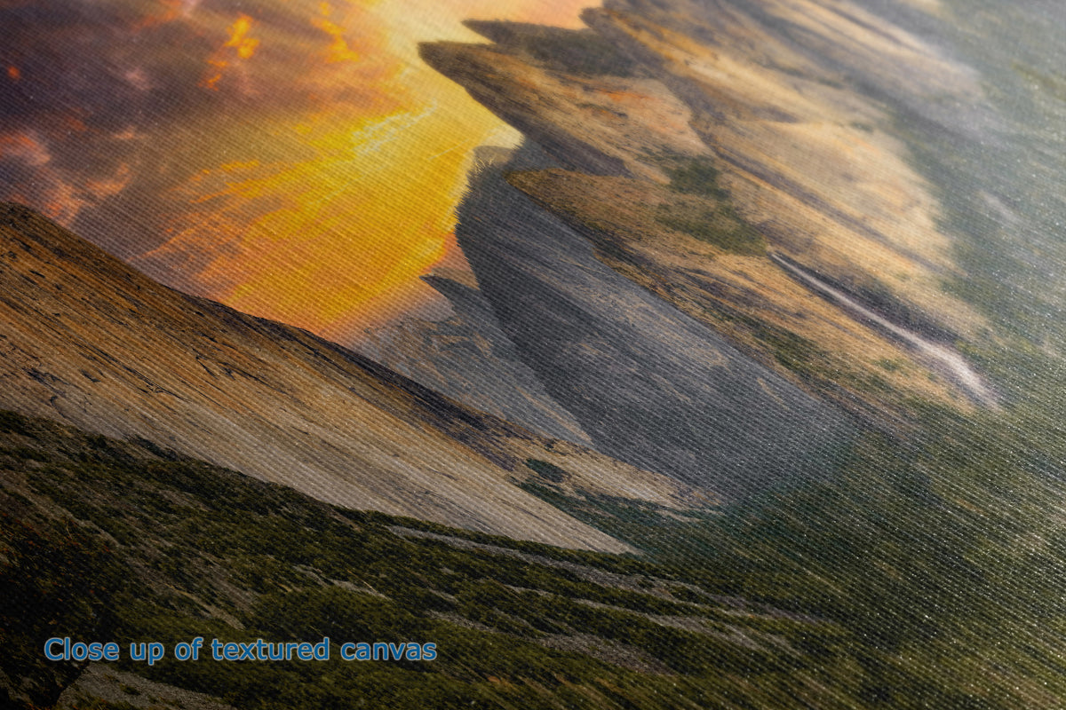 Close-up of the detail and texture of a canvas print depicting a dramatic mountain landscape at sunset, with glowing orange and yellow clouds, dark rocky cliffs, and green-brown terrain. Blue text in the lower-left corner reads 'Close up of textured canvas'.