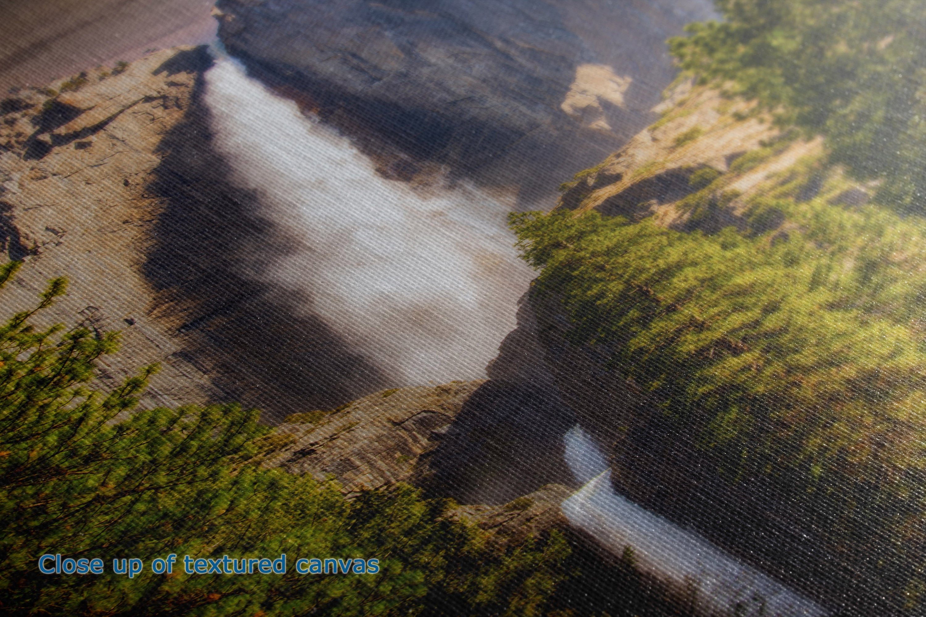 A close-up of a textured canvas print depicting a scenic landscape with a waterfall cascading down a rocky cliff into a river, surrounded by green foliage. A blue text overlay in the bottom left corner reads 'Close up of textured canvas'.