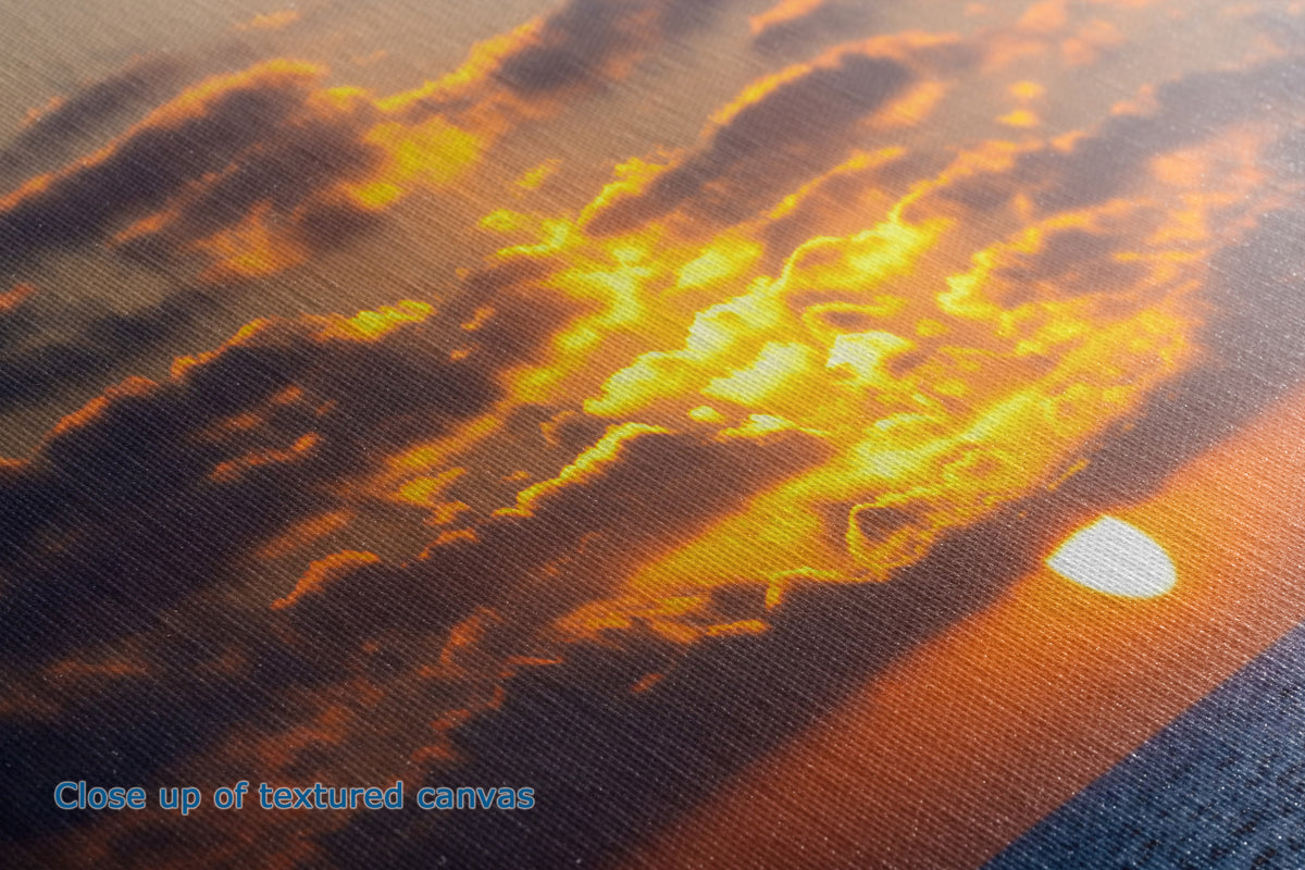 Close-up of a textured canvas print depicting a dramatic sunset sky with glowing yellow and orange clouds, dark brown shadows, and a bright sun near the horizon.