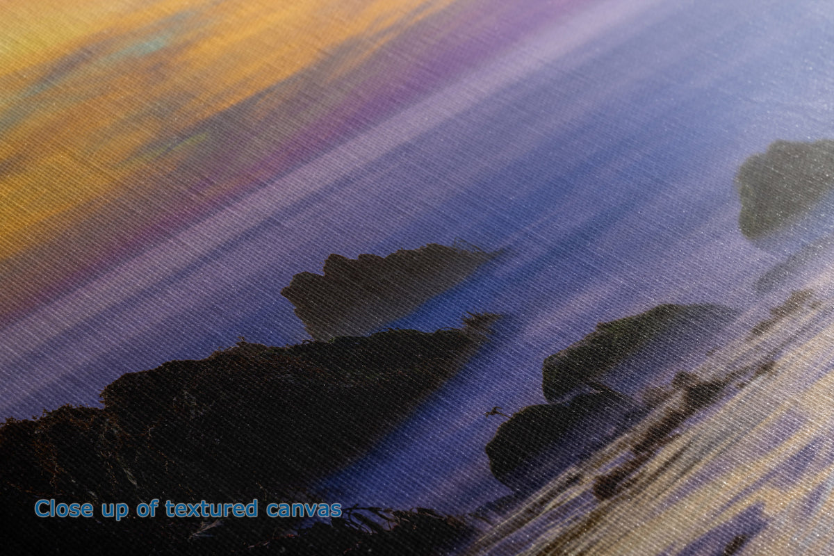 Close-up of a textured canvas print showing a coastal landscape with dark rocky cliffs against a sunset sky with yellow, orange, and purple tones.