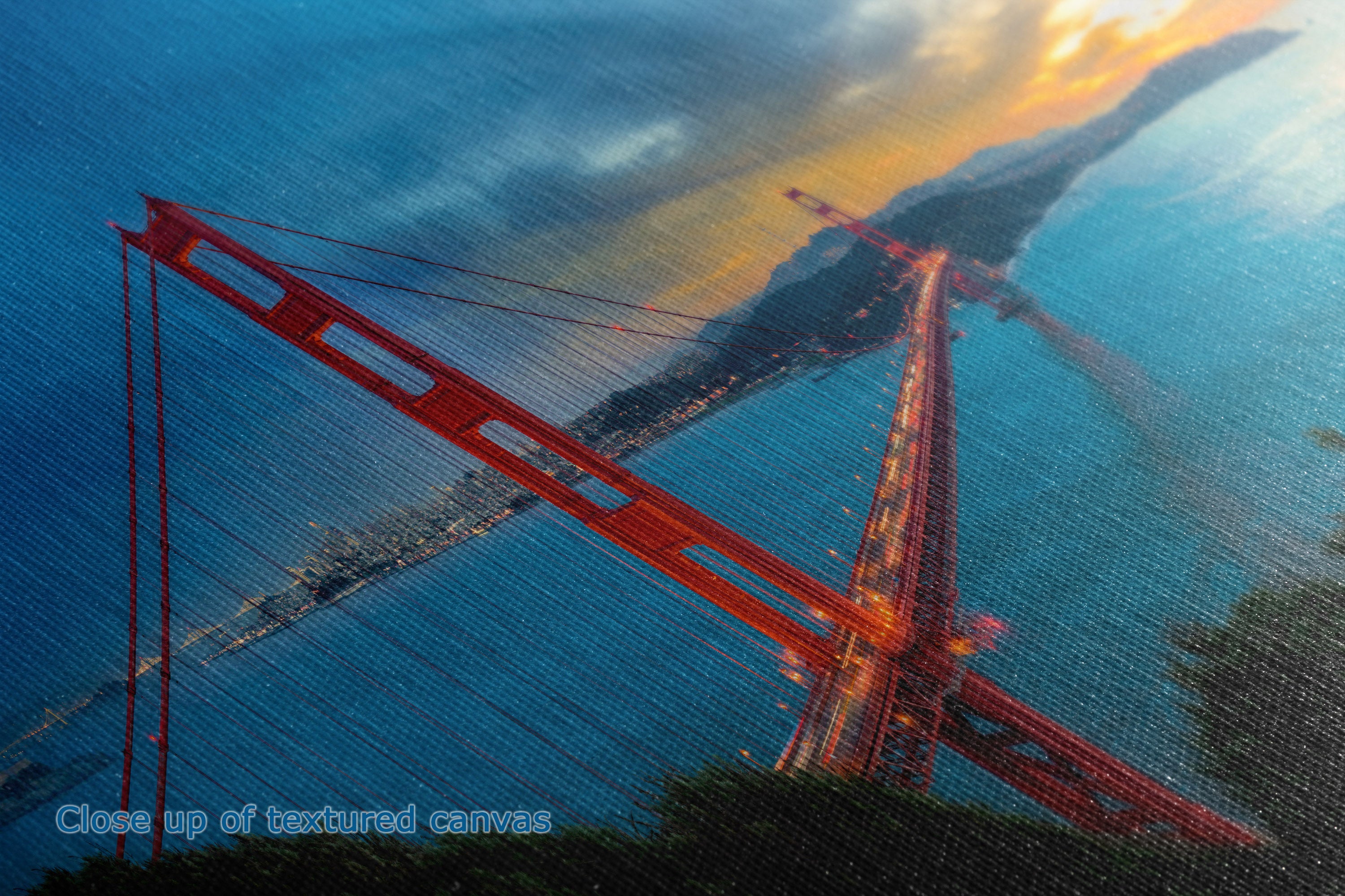 A close-up of a canvas print depicting a dramatic red suspension bridge over blue water under a golden sunset sky, with a faint canvas texture visible.