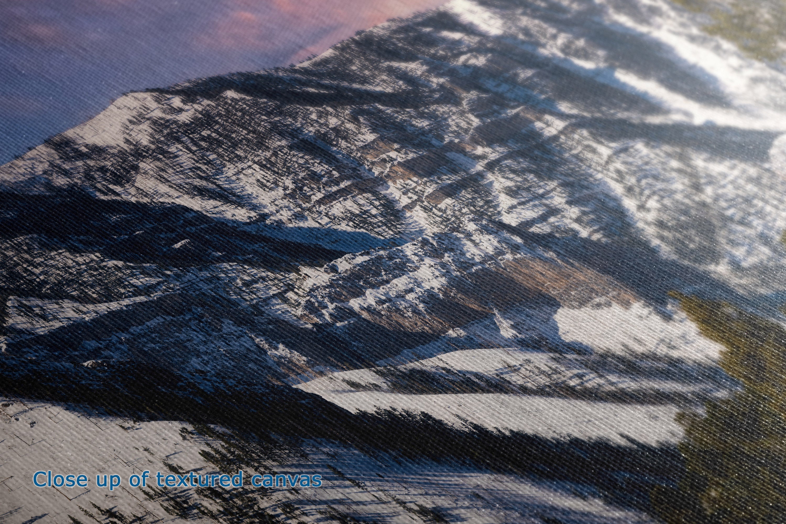 Close-up of a textured canvas print depicting snowy mountain peaks under a pink and blue sky, with blue text in the lower-left corner reading 'Close-up of textured canvas'.