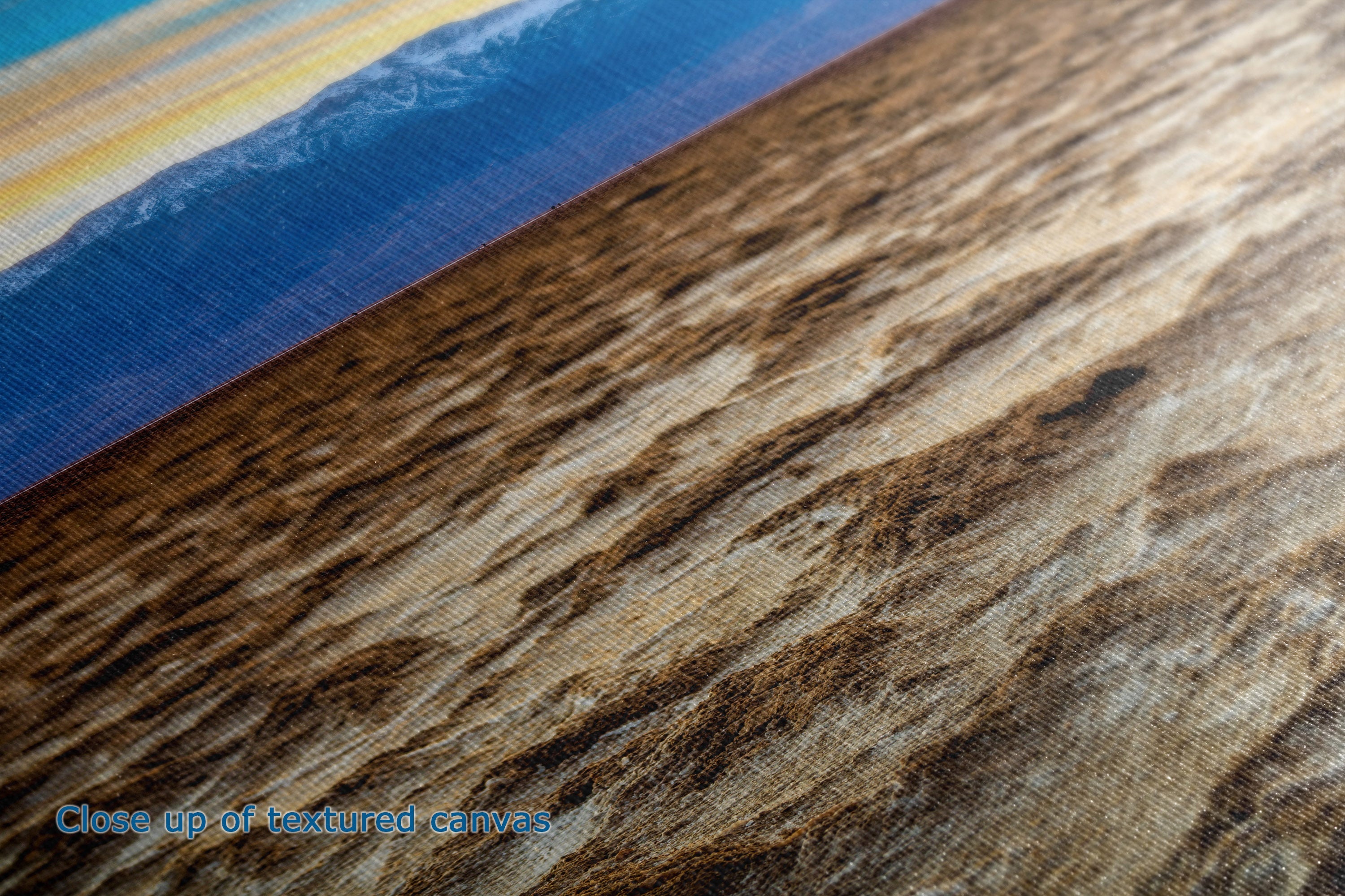 Close-up of a textured canvas print showing a beach scene with blue ocean water and a warm brown sandy shore, with a small blue text overlay reading 'Close up of textured canvas'.
