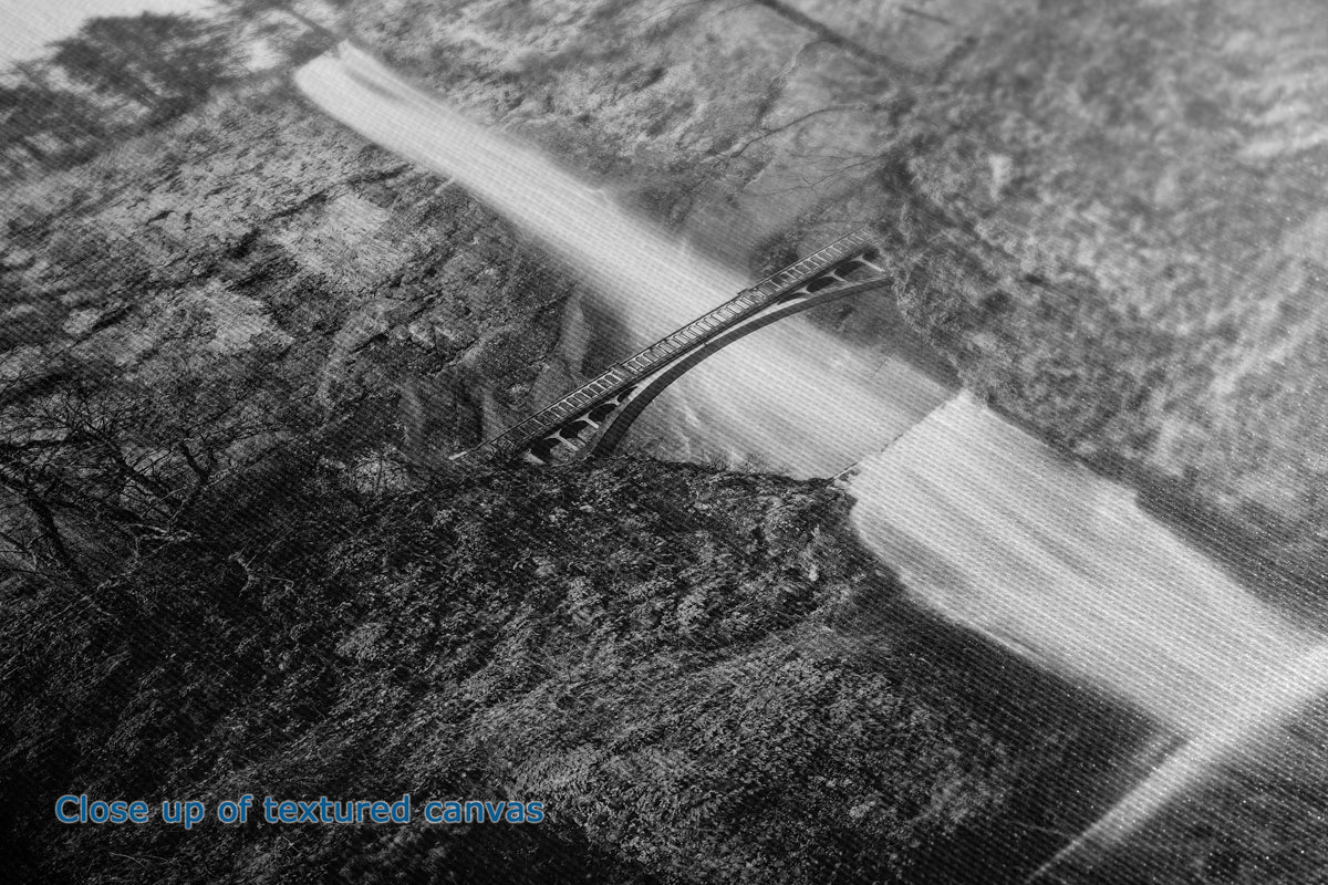 Black and white close-up of a textured canvas print depicting a bridge spanning a rushing waterfall over a rocky landscape, with blue text in the lower-left corner reading 'Close up of textured canvas.'.