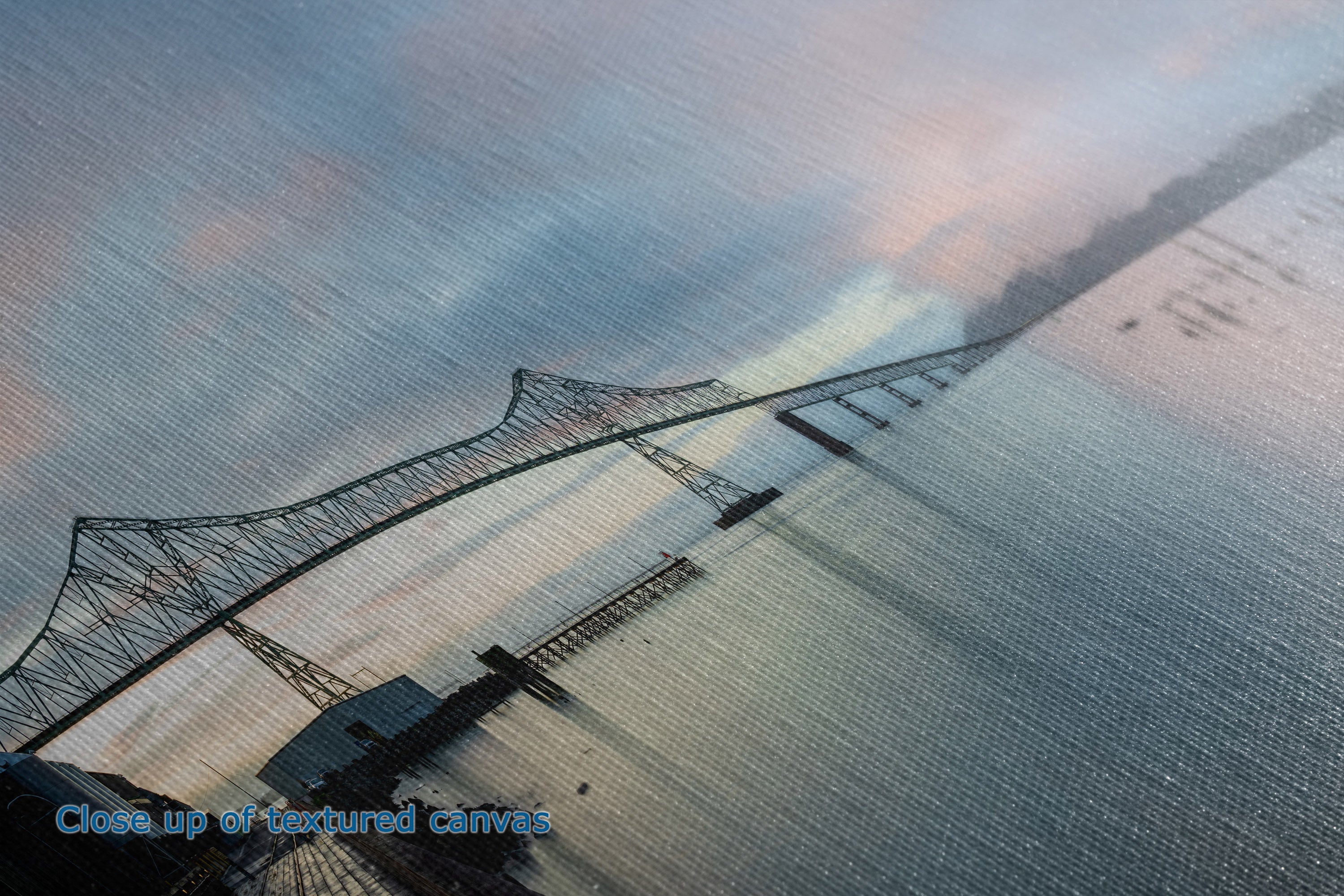 Close-up of a textured canvas print depicting a suspension bridge over water under a cloudy sky at dusk or dawn, with blue-gray tones and soft pink highlights.
