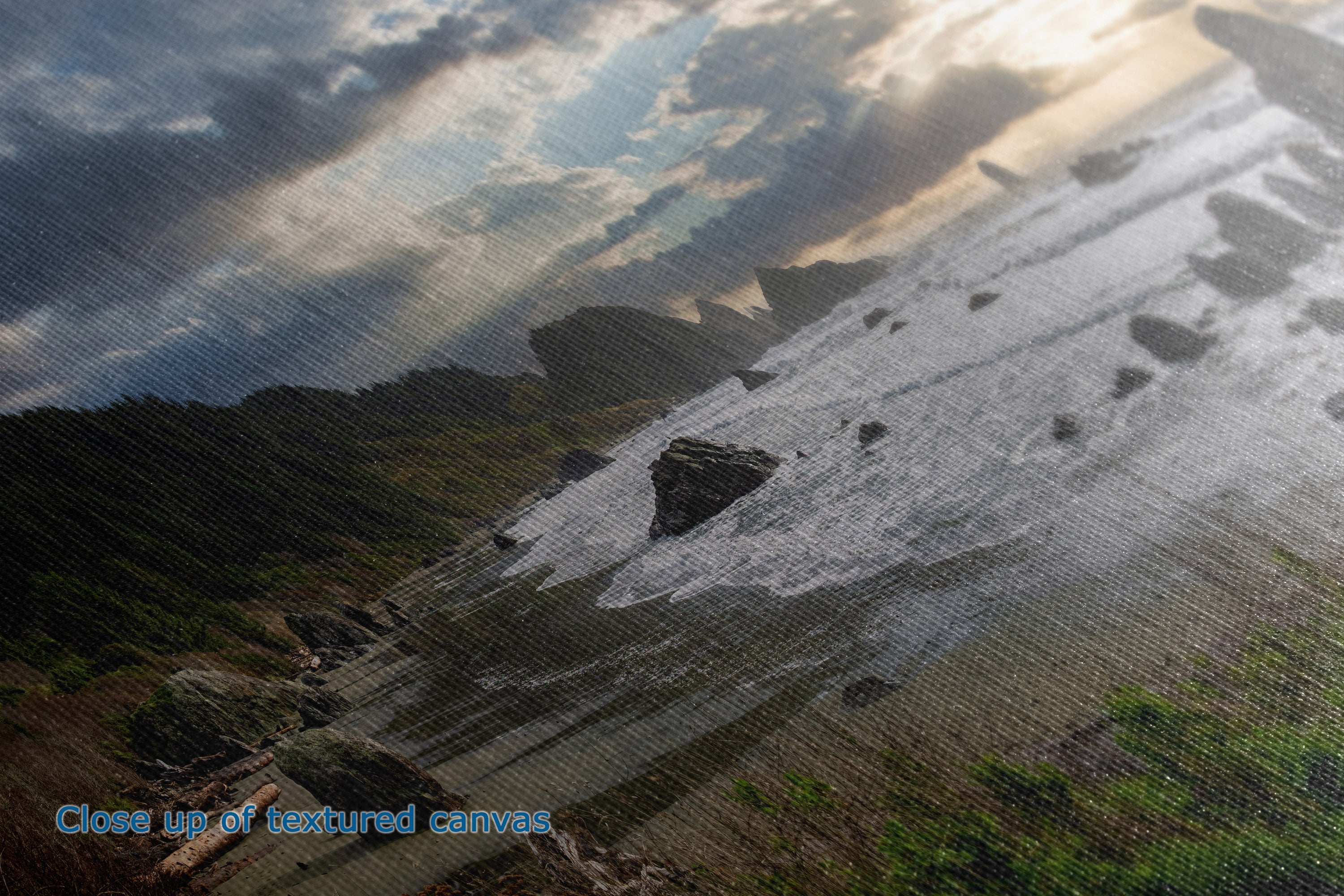 A close-up of a textured canvas print depicting a dramatic coastal landscape with dark rocky cliffs, a sandy shore, and a cloudy sky with sun rays breaking through. Blue text in the bottom-left corner reads 'Close up of textured canvas.