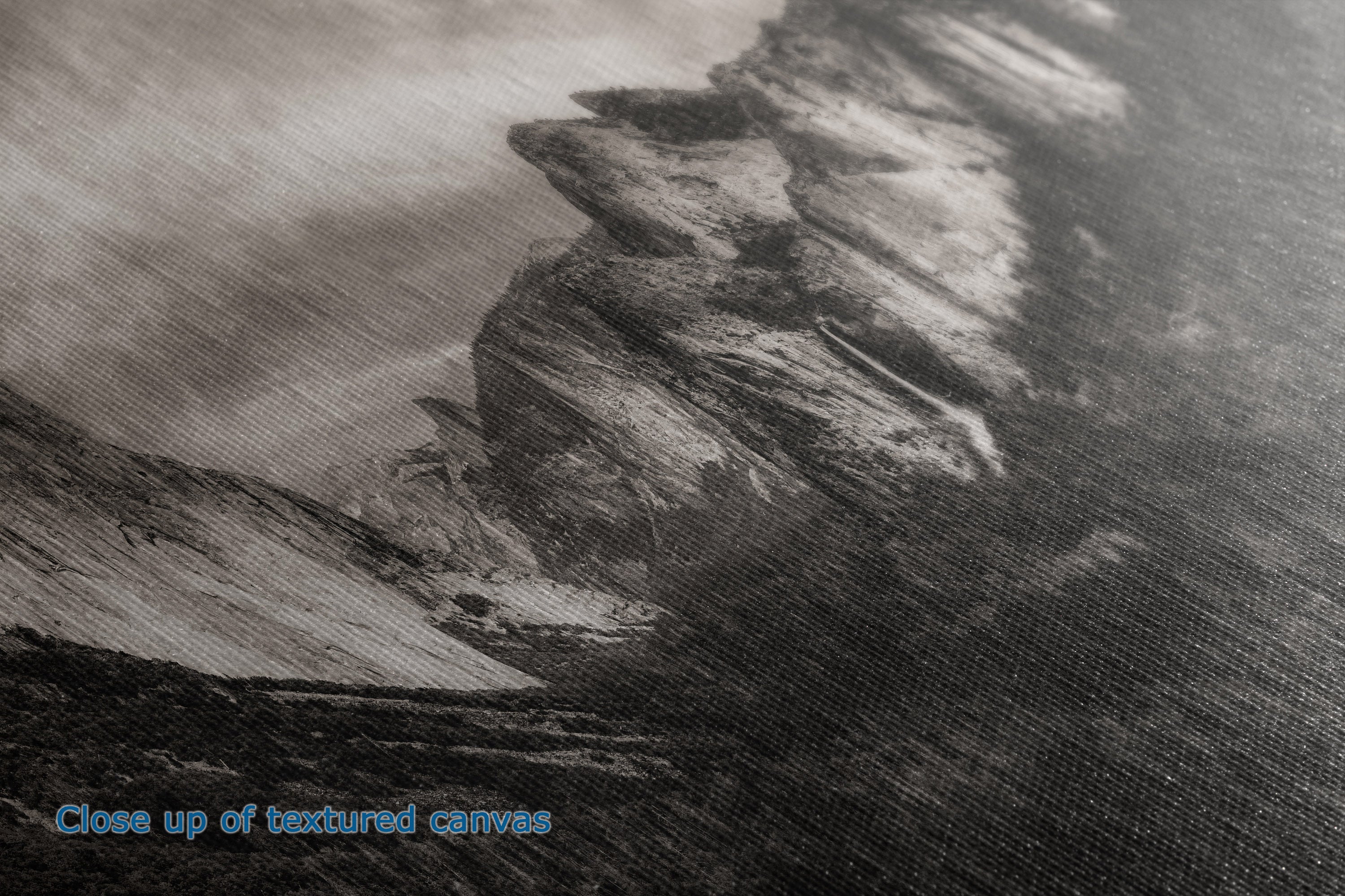 Close-up of a textured canvas print depicting a dramatic rocky cliffside or mountain slope in grayscale tones, with blue text in the lower-left corner reading 'Close up of textured canvas'.