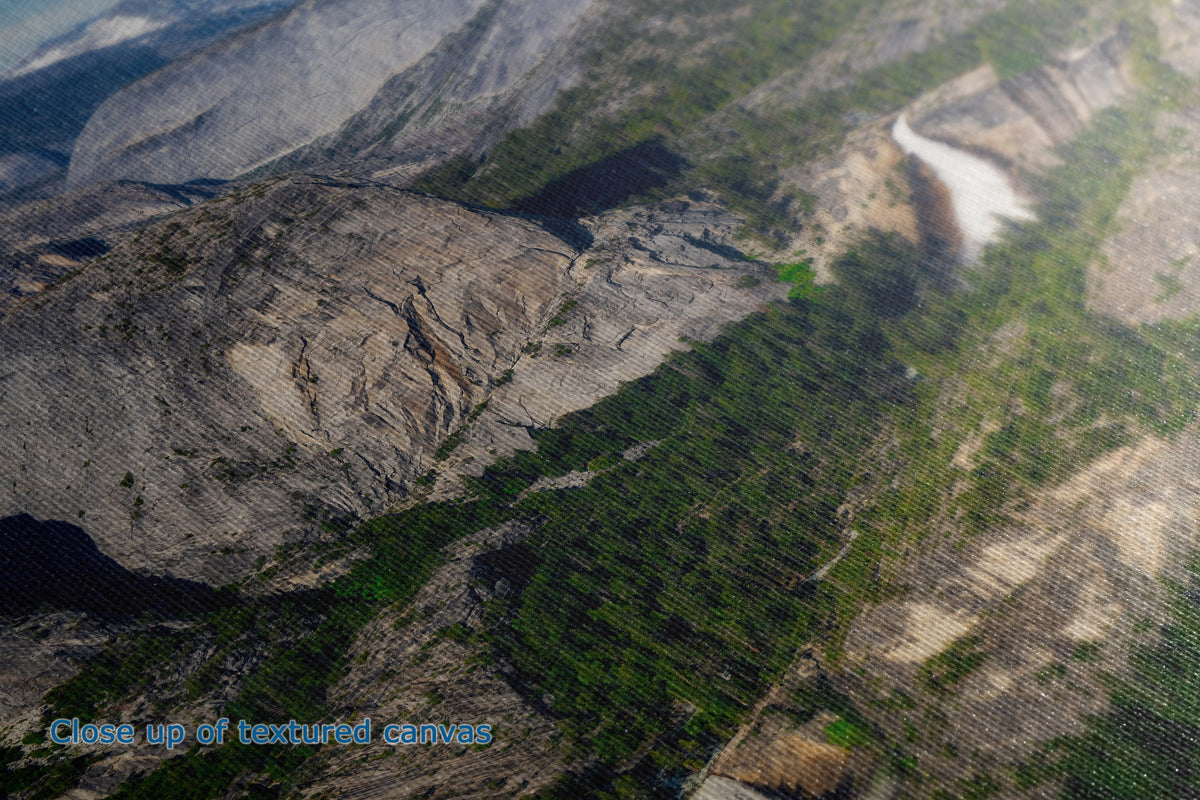 A detailed close up view of a canvas wall art print depicting a mountain landscape with green forested slopes and rocky cliffs, with a blue text overlay reading 'Close up of textured canvas'.