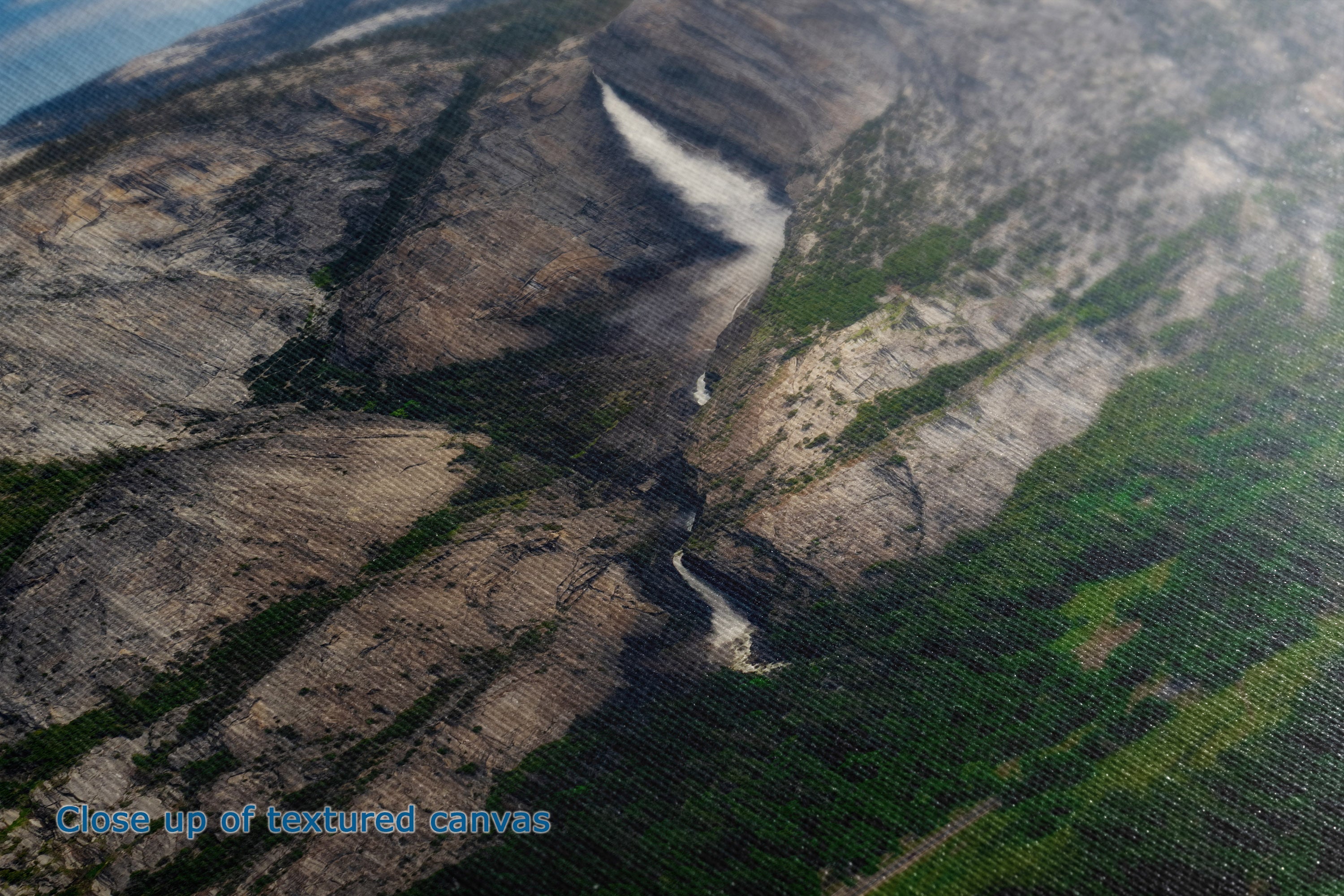 A close up detail and textured view of a canvas wall art print depicting a rugged mountain landscape with a winding river, green forests, and rocky cliffs.