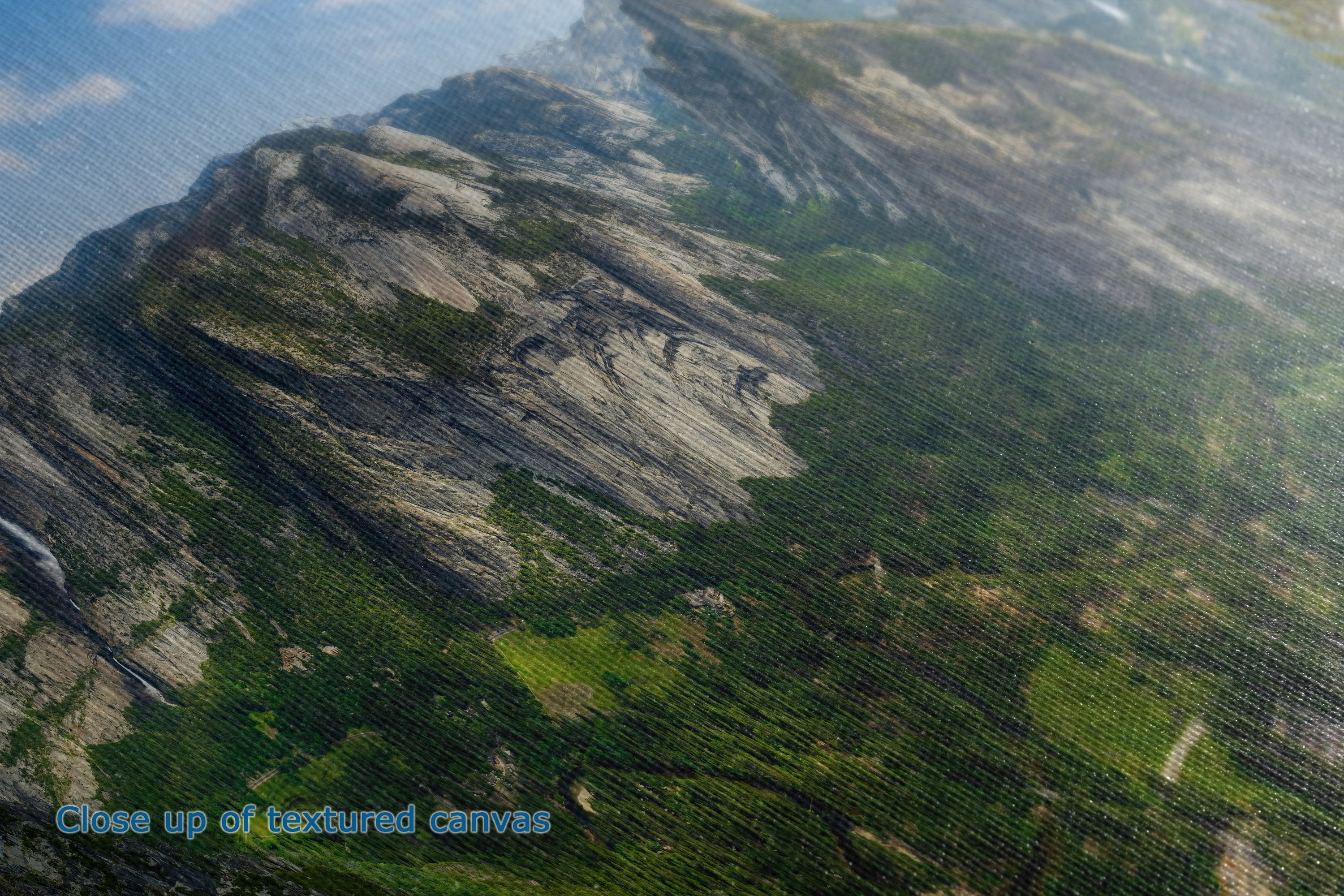 Close-up of the detail and texture of a canvas print depicting a rugged mountain landscape with green forests and rocky cliffs under a blue sky.