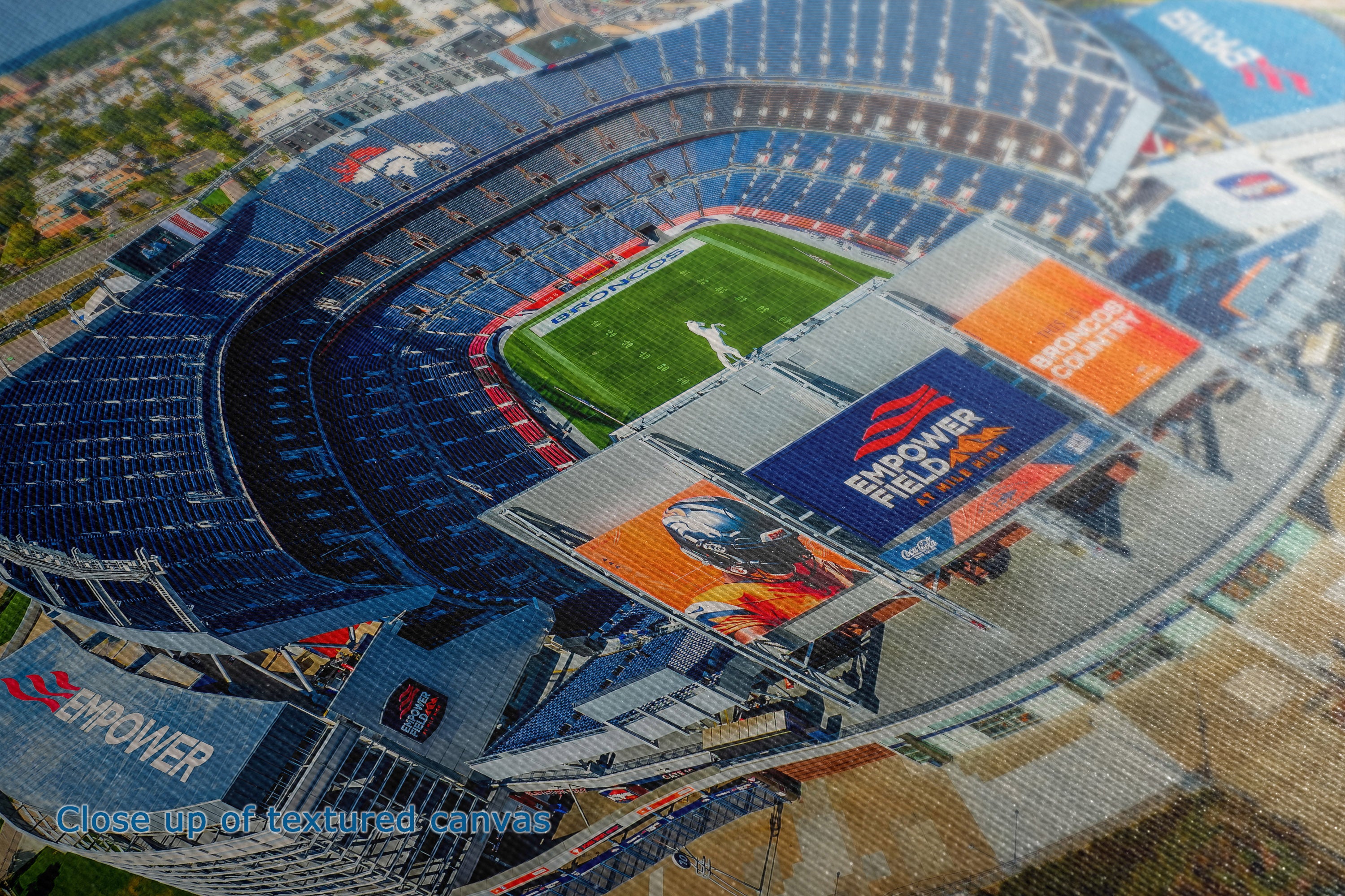A close-up detailed and texture view of a canvas print depicting an aerial view of the Denver Broncos Mile High football stadium, showing the blue seating, green field, and orange advertising boards.