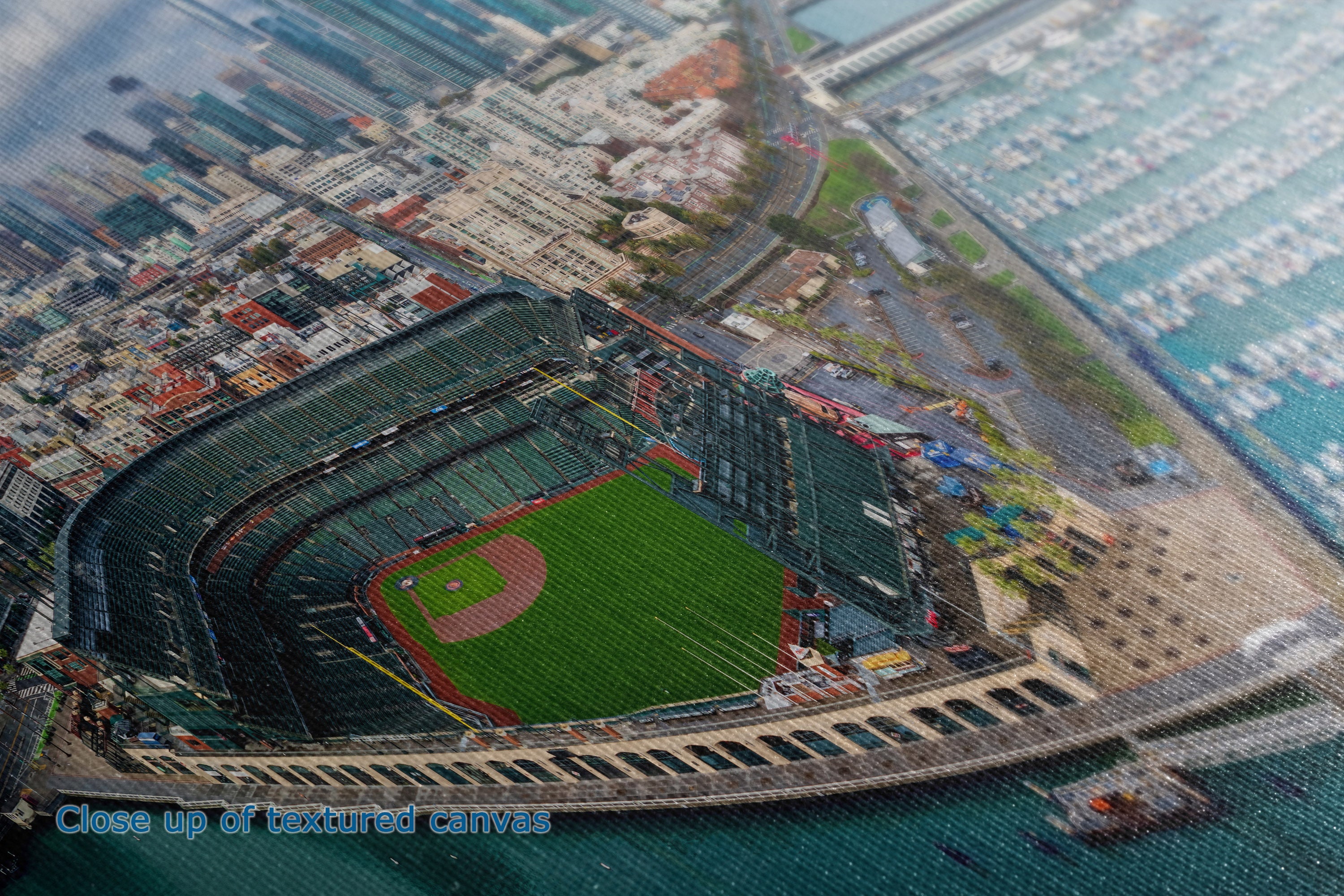 A close-up view of the details and texture of a canvas wall art print depicting an aerial view of a baseball stadium with a green field, dark green seating, an adjacent marina, and surrounding city buildings. A small text overlay at the bottom reads 'Close up of captured canvas.