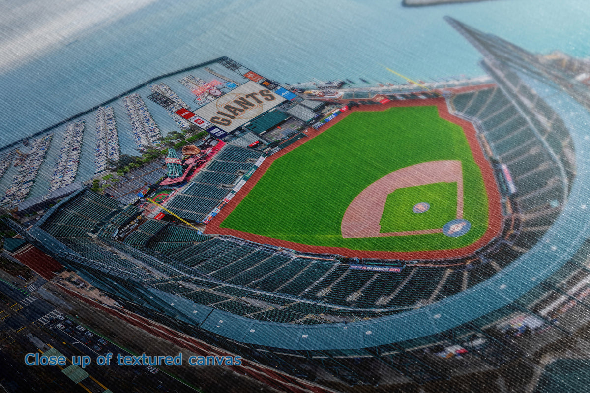 A close-up detail and texture view of a canvas wall art print showing a baseball stadium interior and field, with the word 'GIANTS' visible on the scoreboard. The photo shows the green infield, dirt pitching area, seating tiers, and surrounding blue-gray stadium structure.