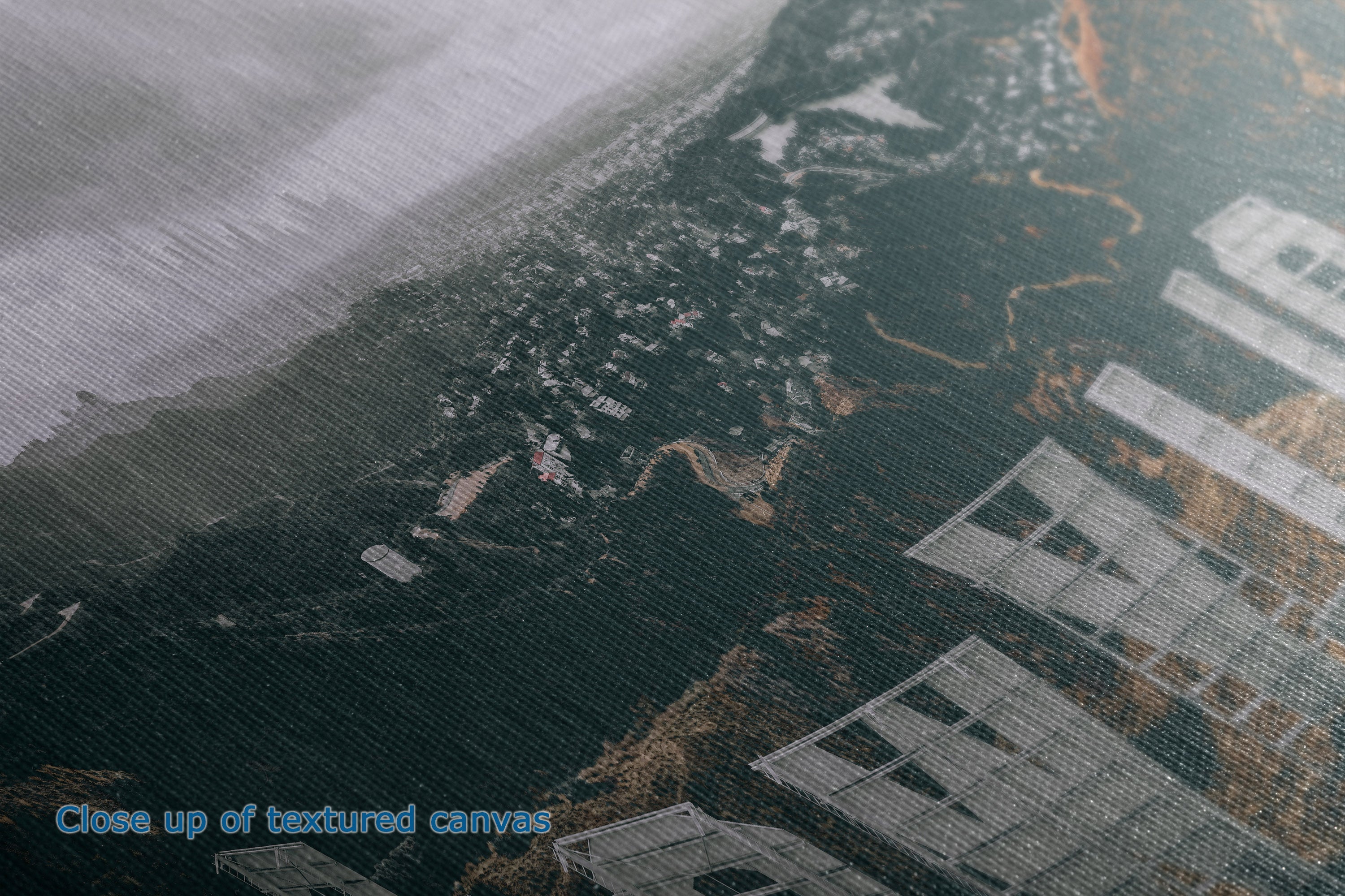 Close-up of a textured canvas print depicting a coastal landscape with dark water, sandy shoreline, and white buildings, with blue text reading 'Close up of textured canvas' in the lower-left corner.