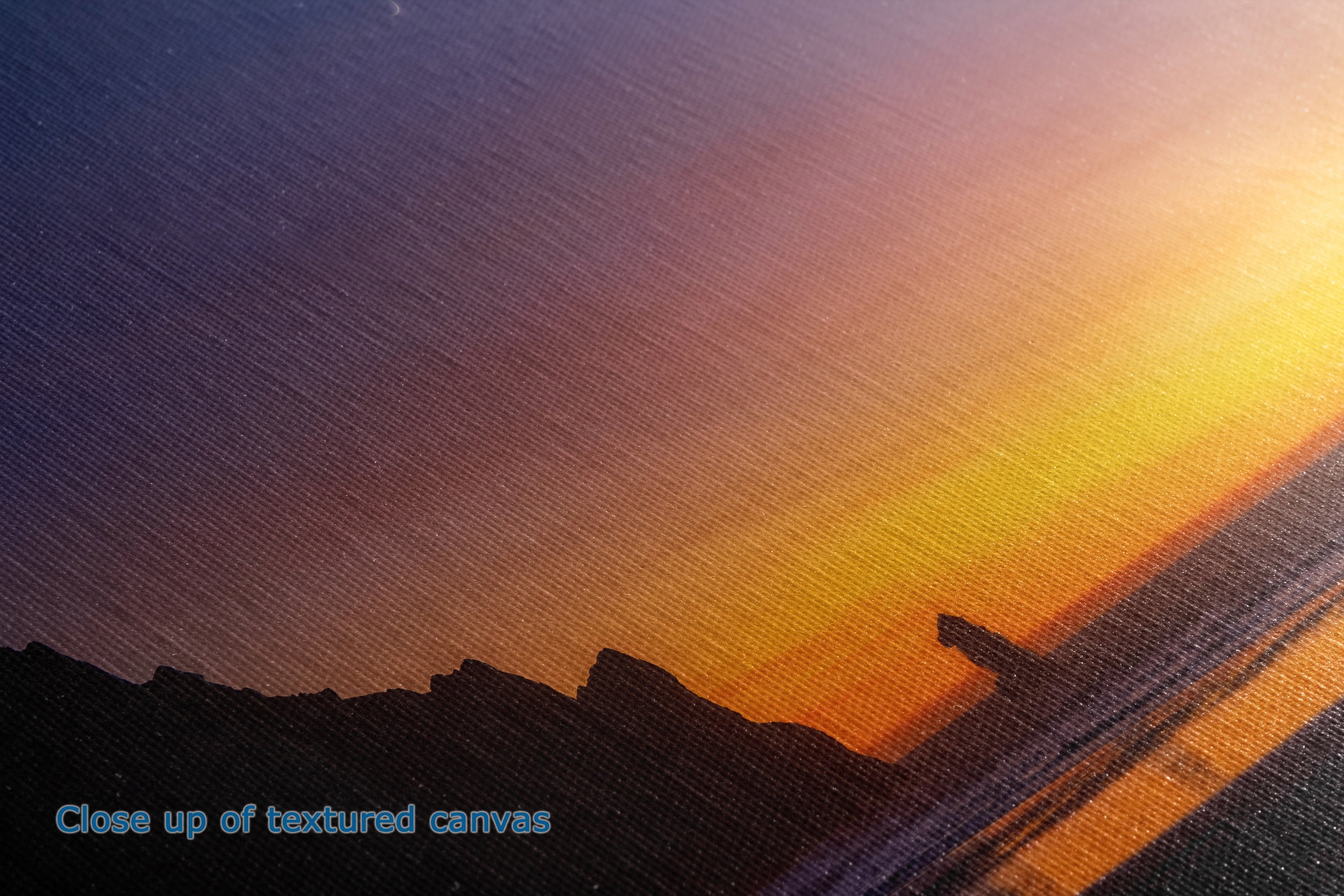 Close-up of a textured canvas print depicting a sunset over a shoreline silhouette, with a blue text overlay reading 'Close up of textured canvas' in the lower-left corner.