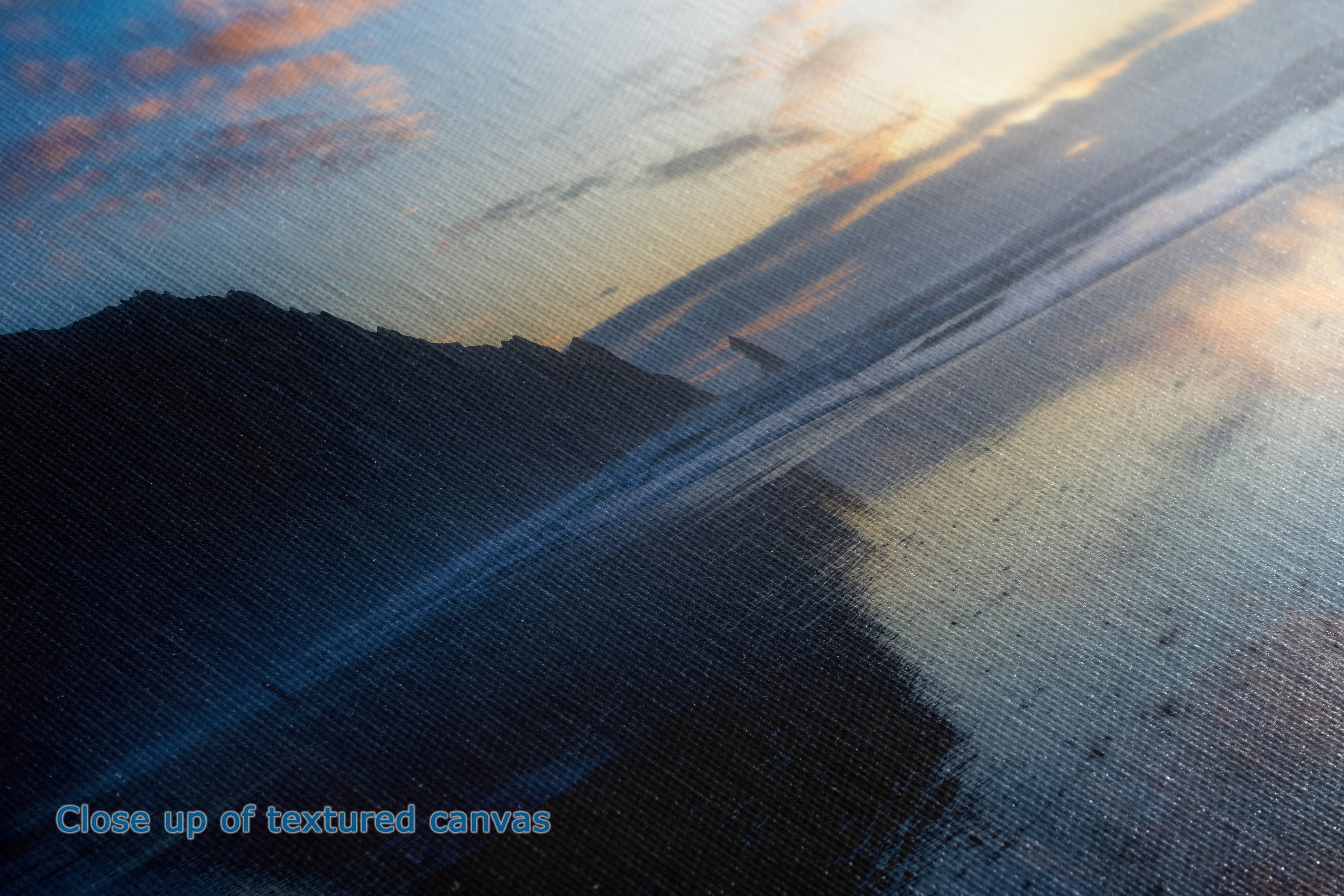 Close-up of a textured canvas print depicting a sunset over water with dark silhouetted landforms, warm orange and yellow light in the sky, and blue-gray tones reflecting across the water. Small blue text in the lower-left corner reads 'Close up of textured canvas'.