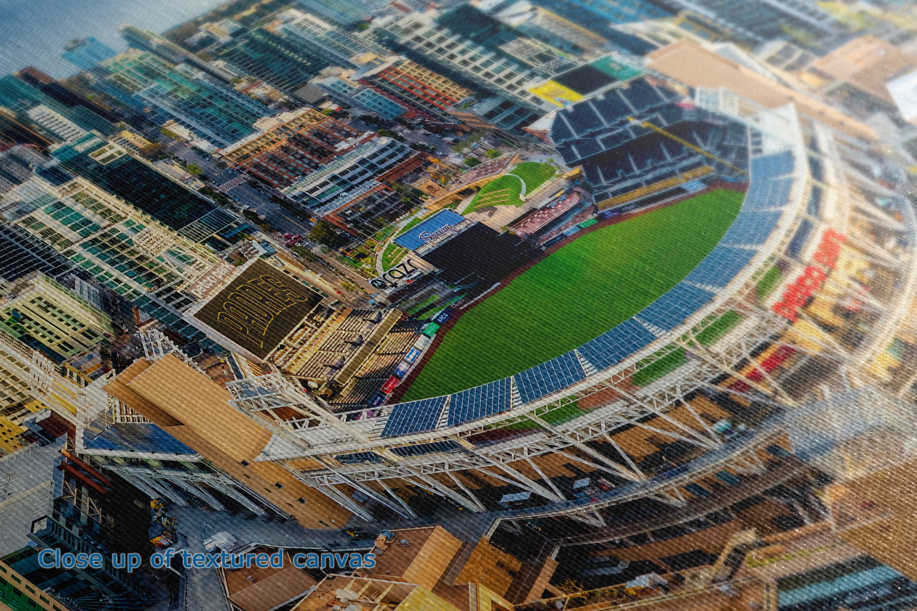 A close-up of the detail and texture of a canvas print depicting an aerial view of a stadium surrounded by dense urban buildings, with a blue text overlay in the bottom-left corner reading 'Close up of textured canvas'.