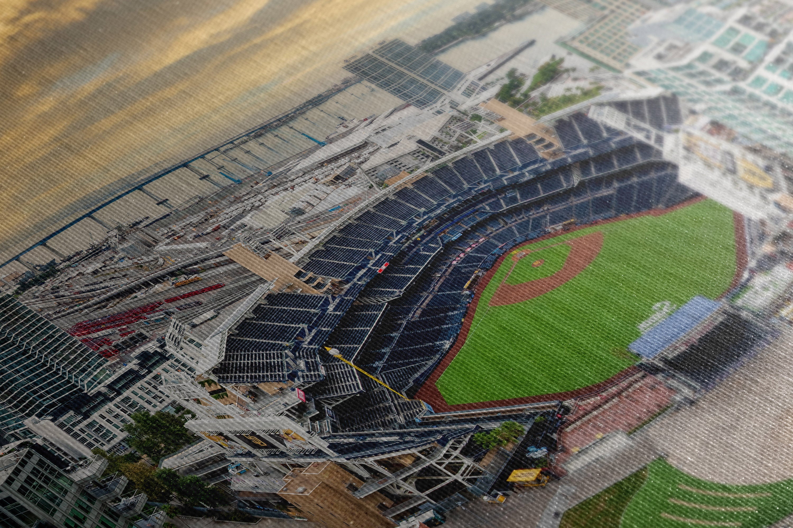 A close-up of a printed or textured baseball stadium scene, showing the field, stands, surrounding buildings, and sky in a detailed overhead view.