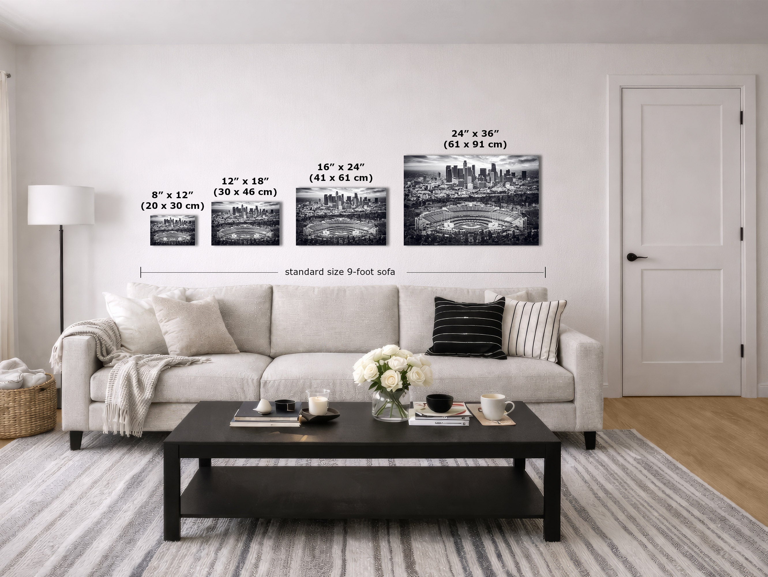 A staged living room showing four framed black and white Los Angeles Dodgers Stadium artwork cityscape prints of mounted on a white wall above a light sofa. The prints are labeled with dimensions: 8" x 12" (20 x 30 cm), 12" x 18" (30 x 46 cm), 16" x 24" (41 x 61 cm), and 24" x 28" (61 x 91 cm), with a sofa width reference below.