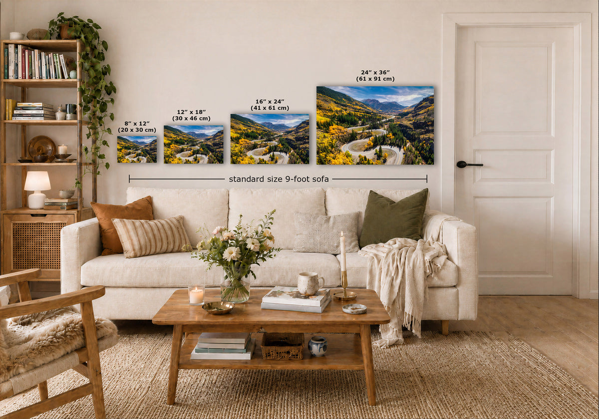 Four framed landscape art prints of a mountain valley with yellow wildflowers are displayed on a white wall above a beige sofa in a styled living room. The prints show different sizes with dimension labels, and the text 'standard size 9-foot sofa' is written below them.