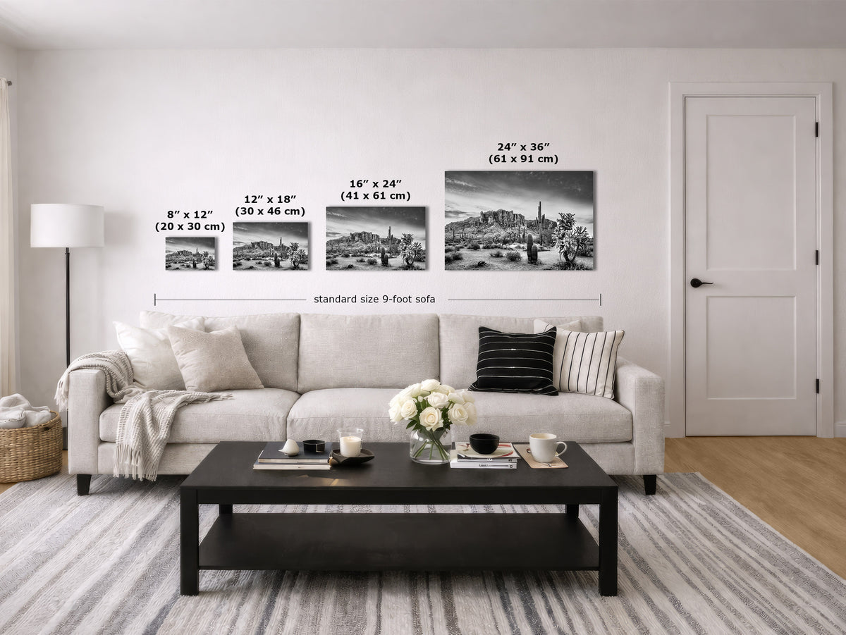 A staged living room showing four black-and-white wall art prints above a light sofa, with size labels and measurements for each print. The room includes a coffee table, rug, lamp, and door.