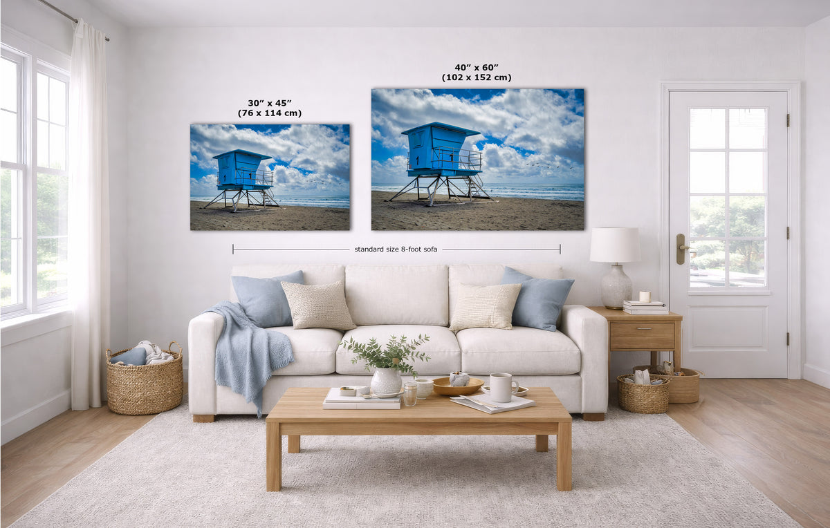 A styled living room showing two canvas wall art prints above a white sofa. The prints depict a blue lifeguard hut on a sandy beach under a cloudy sky.