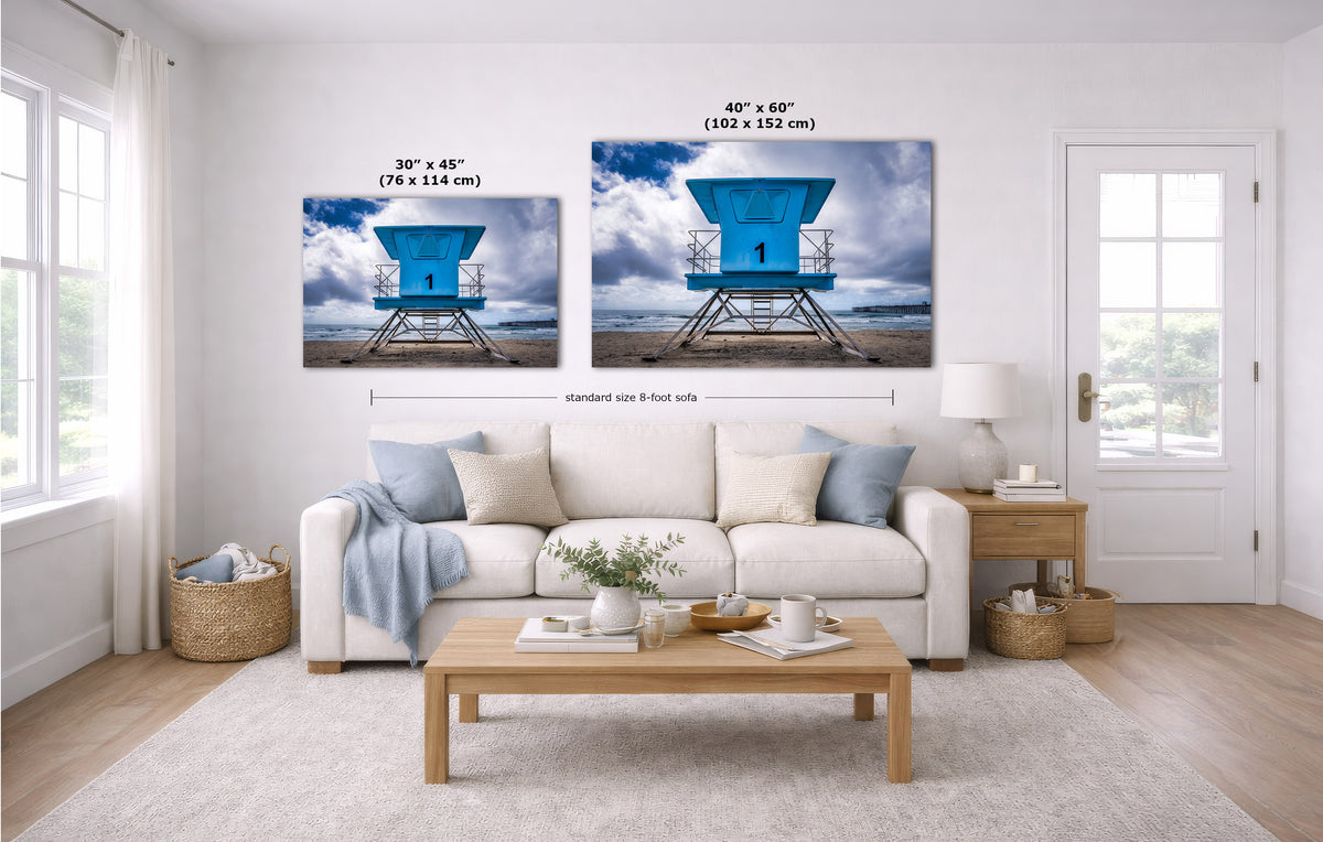 A staged living room shows two vertical wall art prints above a white sofa. Each print depicts a bright blue lifeguard tower scene with clouds and water, and size text is overlaid above each frame.