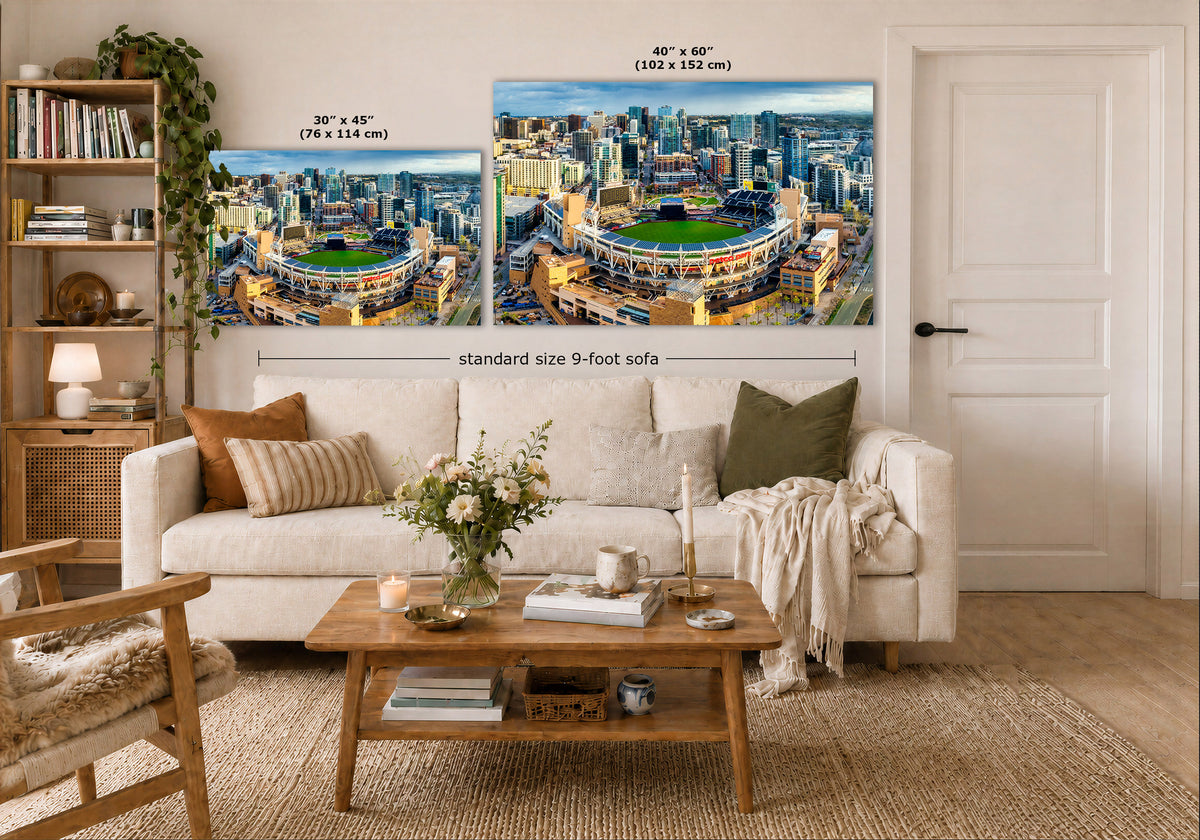 Two large wall art prints showing stadium cityscapes are displayed above a white sofa in a styled living room. The image includes size callouts for the prints: 30 x 45 inches on the left and 40 x 60 inches on the right, with a note reading 'standard size 9-foot sofa'.