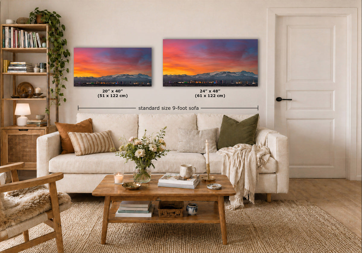 Two framed wall art prints showing a colorful sunset mountain landscape hang above a white sofa in a styled living room. Black dimension lines and text indicate the sizes of both prints, with the left print labeled 20" x 40" (51 x 122 cm) and the right print labeled 24" x 48" (61 x 126 cm), and the sofa labeled standard size 9-foot sofa.