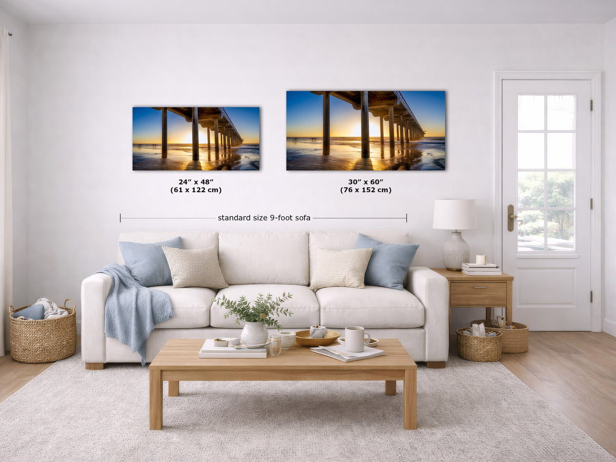 A styled living room showing two framed wall art prints above a white sofa. The prints depict a pier at sunset over water, with size labels and a dimension line indicating standard size 9-foot sofa spacing.
