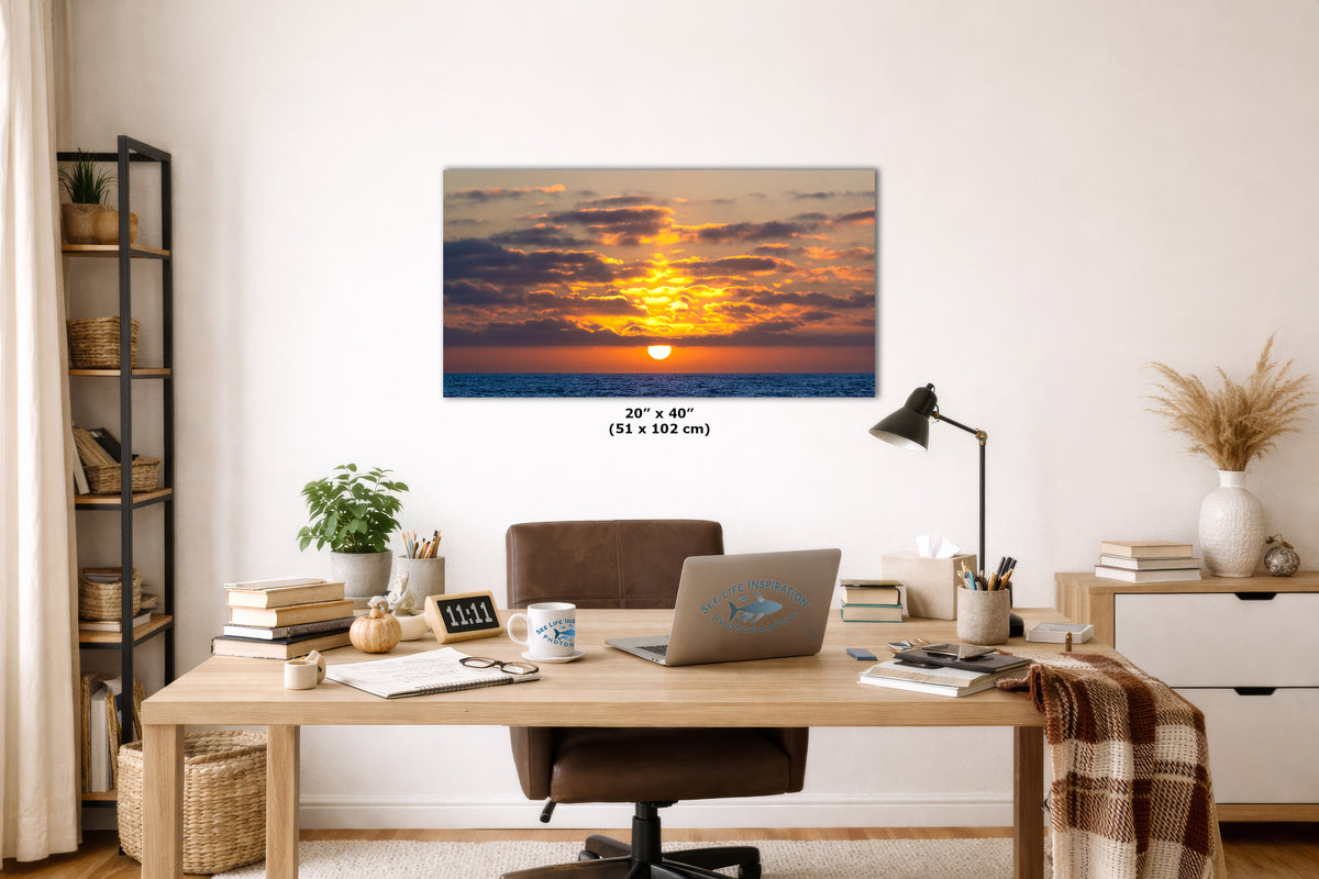 A rectangular wall art print showing a colorful sunset over the ocean, displayed above a wooden desk in a styled home office interior. The print includes size text reading 20 x 40 inches (51 x 102 cm) below the artwork.