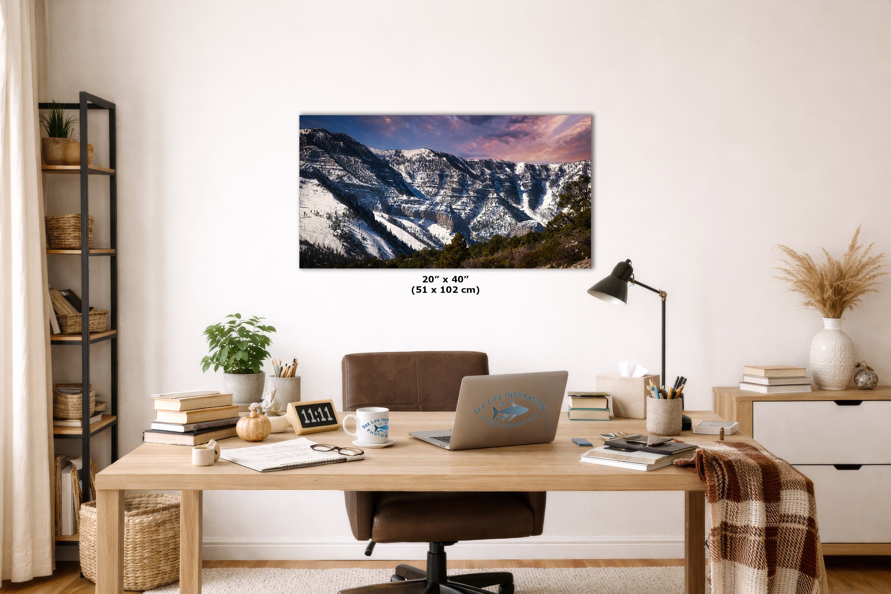 A rectangular wall art print showing a snowy mountain landscape with a pink-purple sky, displayed above a wooden desk in a styled home office. The print includes size text reading 20 x 40 inches (51 x 102 cm).