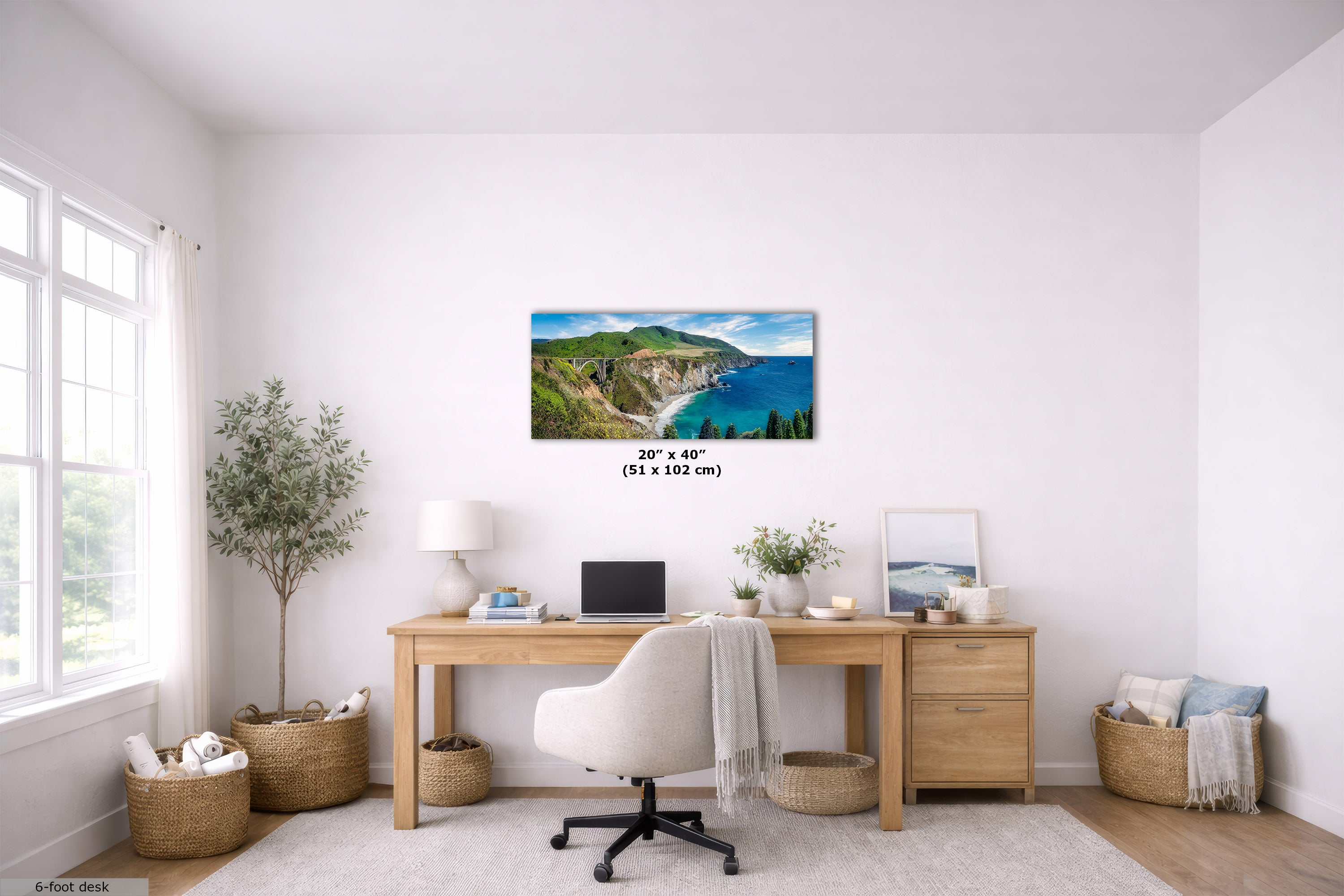 A wall art print showing a coastal landscape with green cliffs and blue ocean, displayed above a wooden desk in a bright, modern home office. The print includes size text reading 20 x 40 (51 x 102 cm) below it.