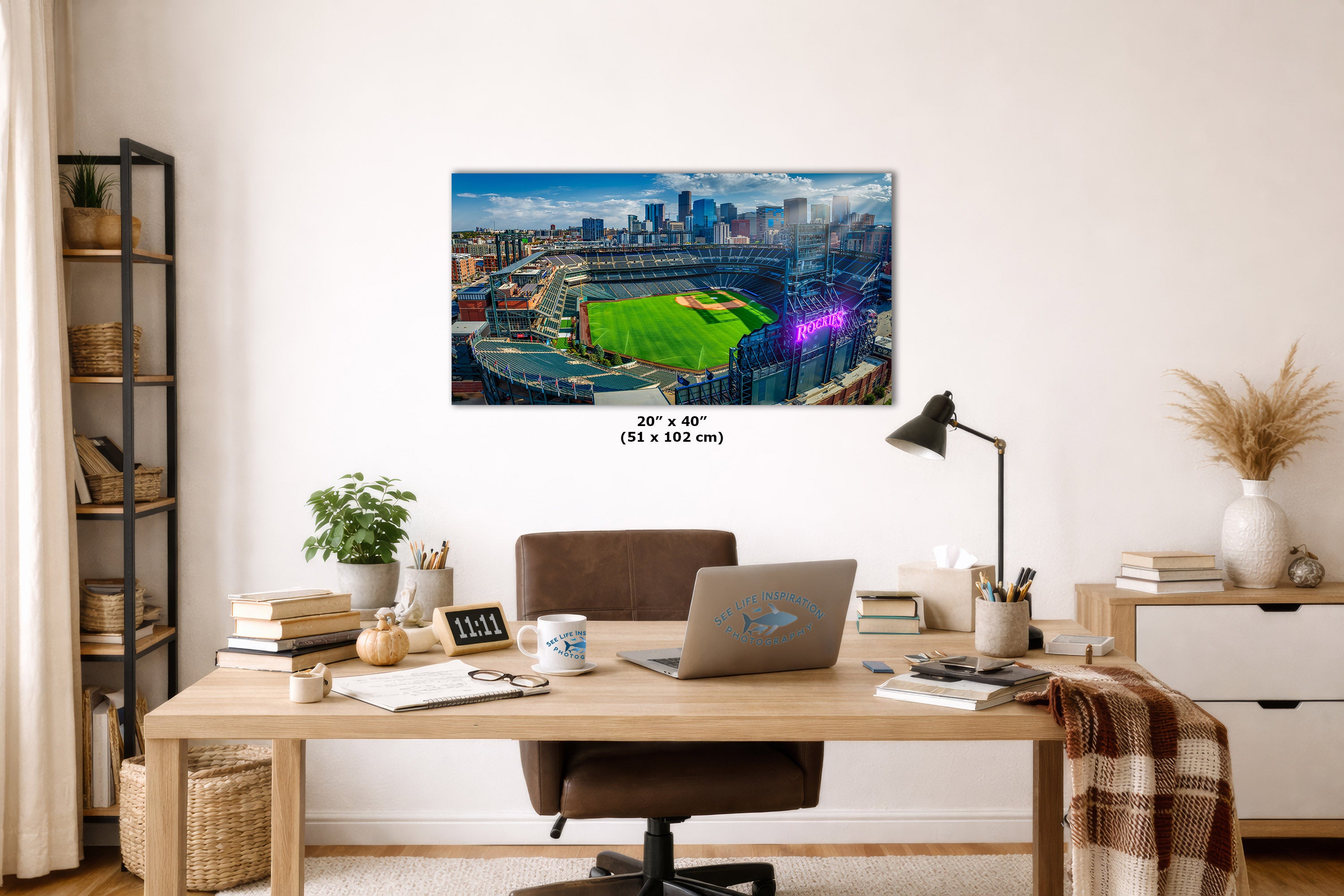 A wall art print showing a baseball stadium scene hangs above a wooden desk in a styled home office. The print depicts a green field, stadium seating, and a city skyline under a blue sky.