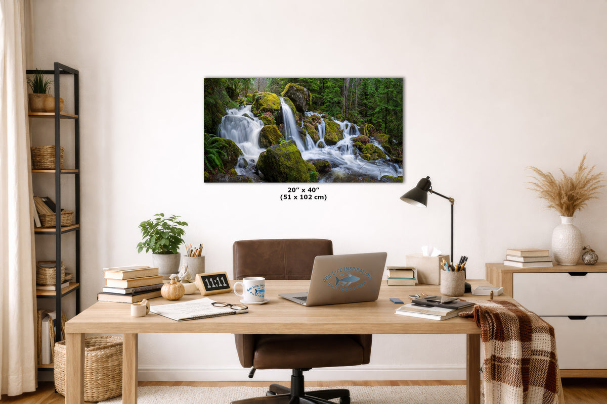 A framed wall art print showing a lush green forest waterfall, displayed in a modern home office setting above a wooden desk with a laptop, lamp, and books. The print includes size markings of 20" x 40" (51 x 102 cm) at the bottom.