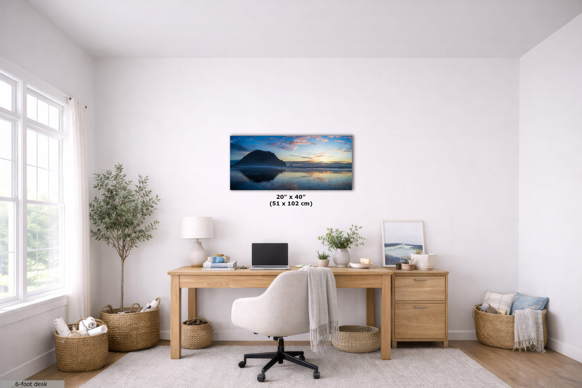 A panoramic landscape art print of a mountain lake at sunset is displayed on a white wall in a modern, bright home office setting with a wooden desk, white chair, and plants.