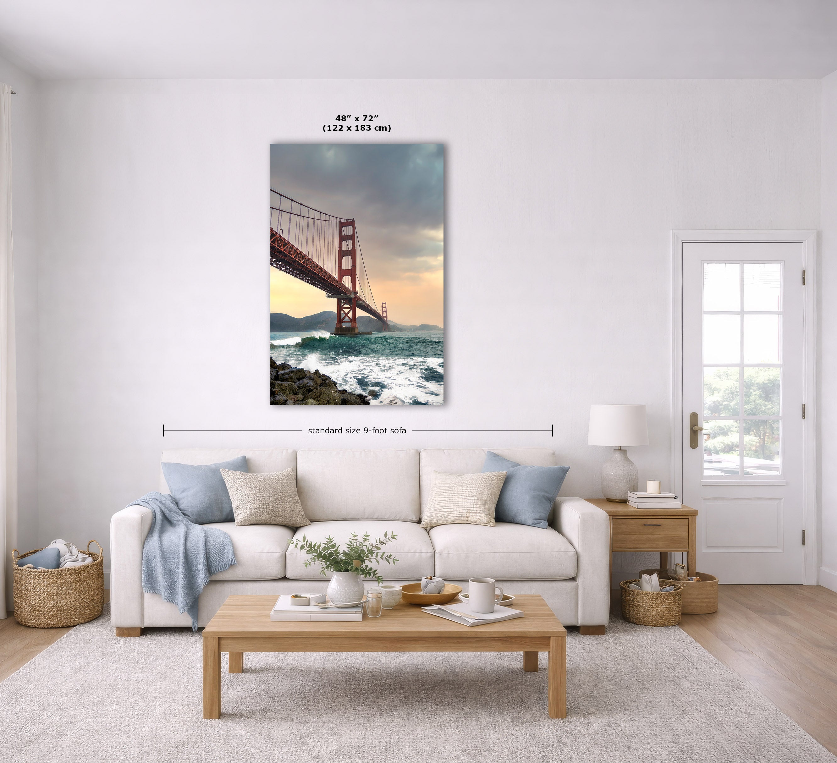 A framed wall art print of the Golden Gate Bridge is displayed above a white sofa in a bright, modern living room. The print shows the iconic orange-red bridge over churning ocean waves at sunset.