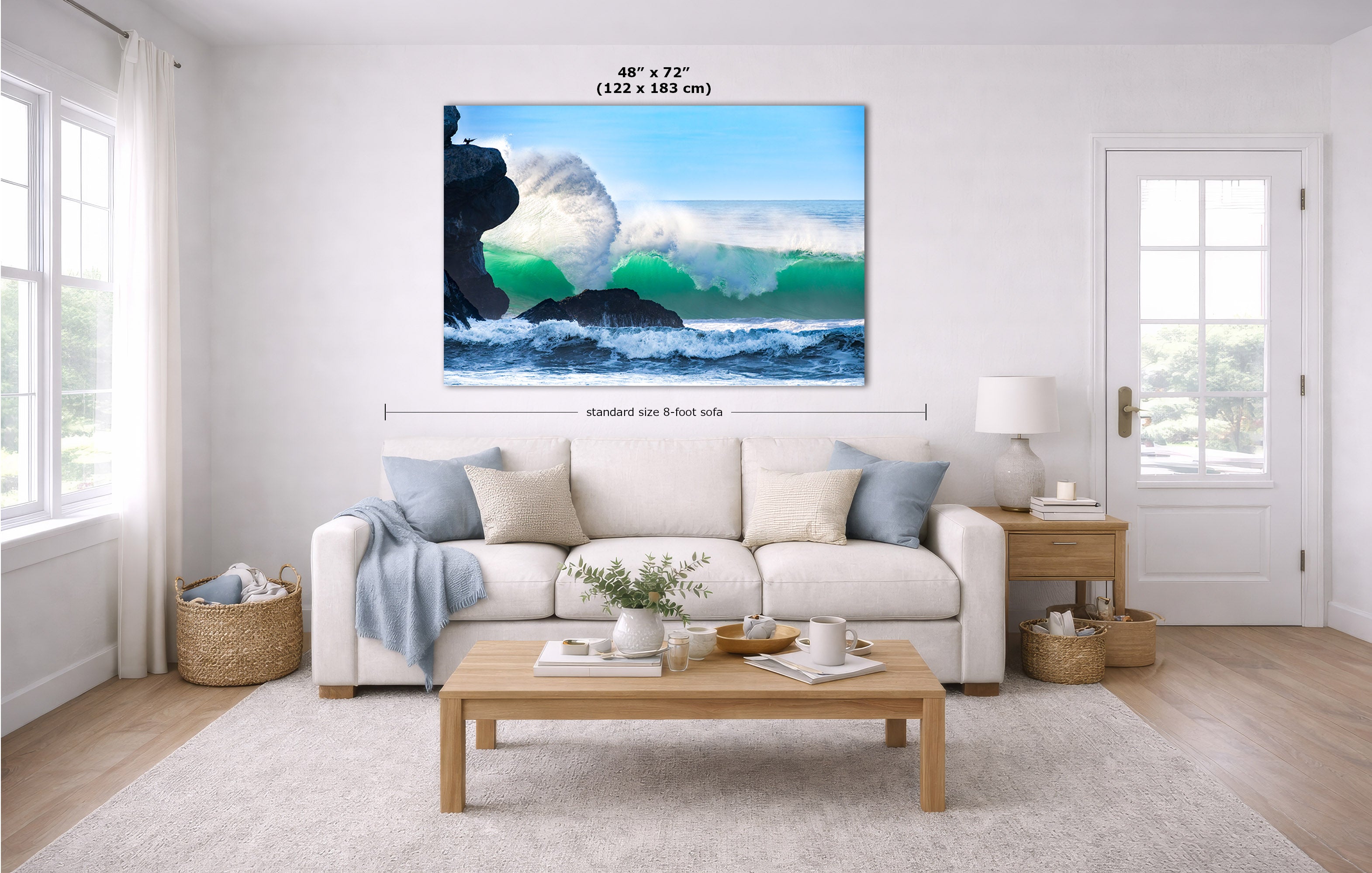 A large rectangular wall art print showing a dramatic ocean wave crashing over dark rocks, displayed above a white sofa in a bright modern living room. Small dimension labels are overlaid near the top and center of the artwork.